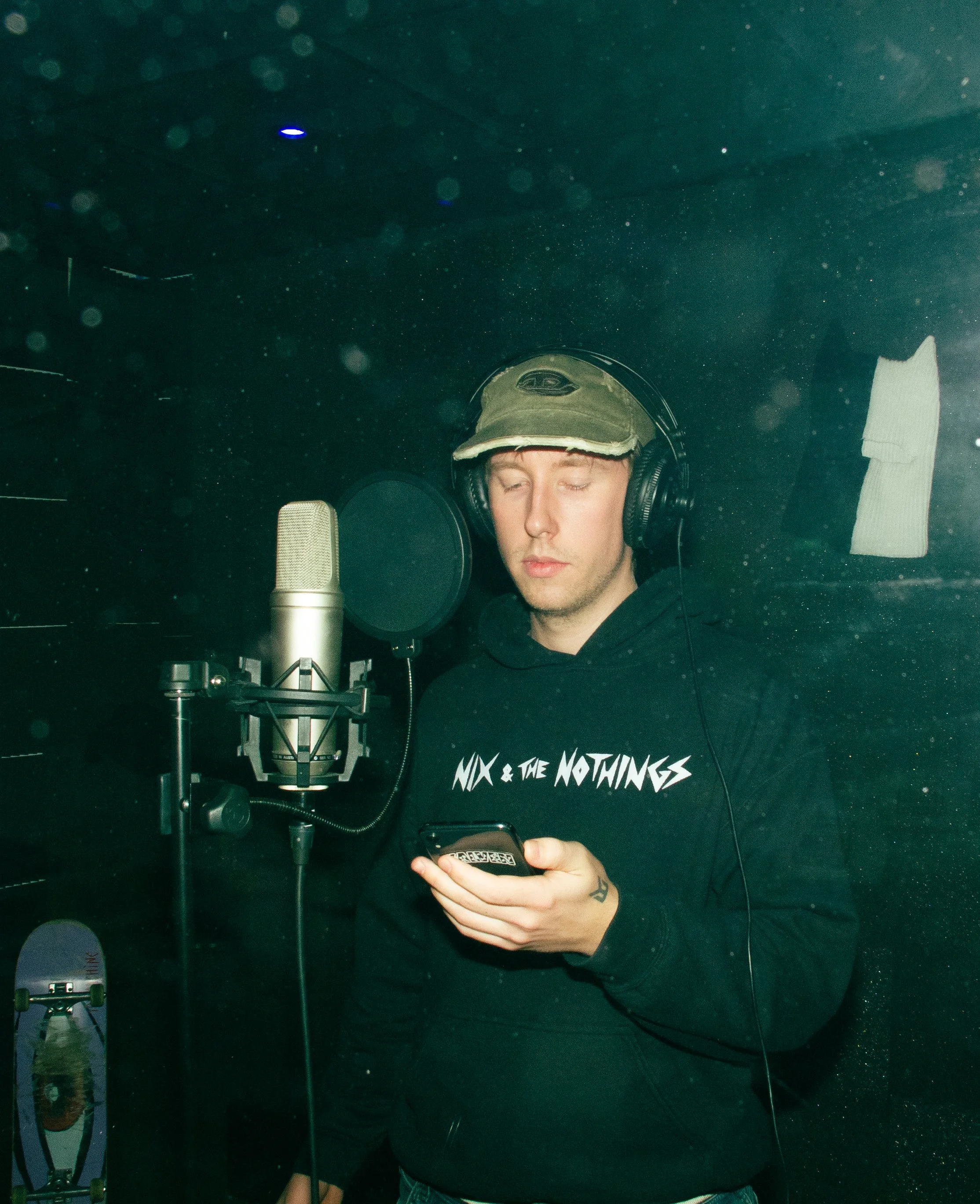 A young man in a recording studio wearing headphones, a cap, and a black hoodie with the text 'NIX & THE NOTINGS', standing near a microphone while looking at his phone.