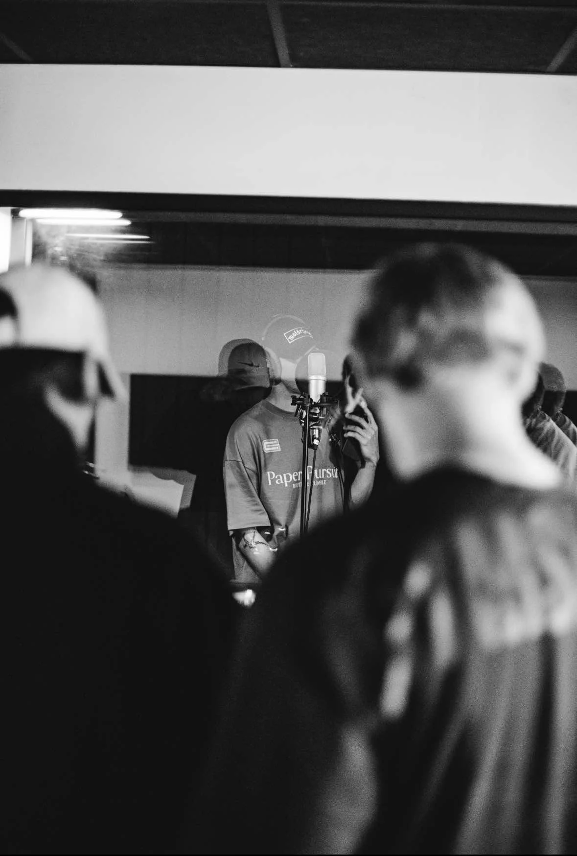 Black and white photo of a recording studio with a singer in front of a microphone, surrounded by blurred figures of people in the foreground.