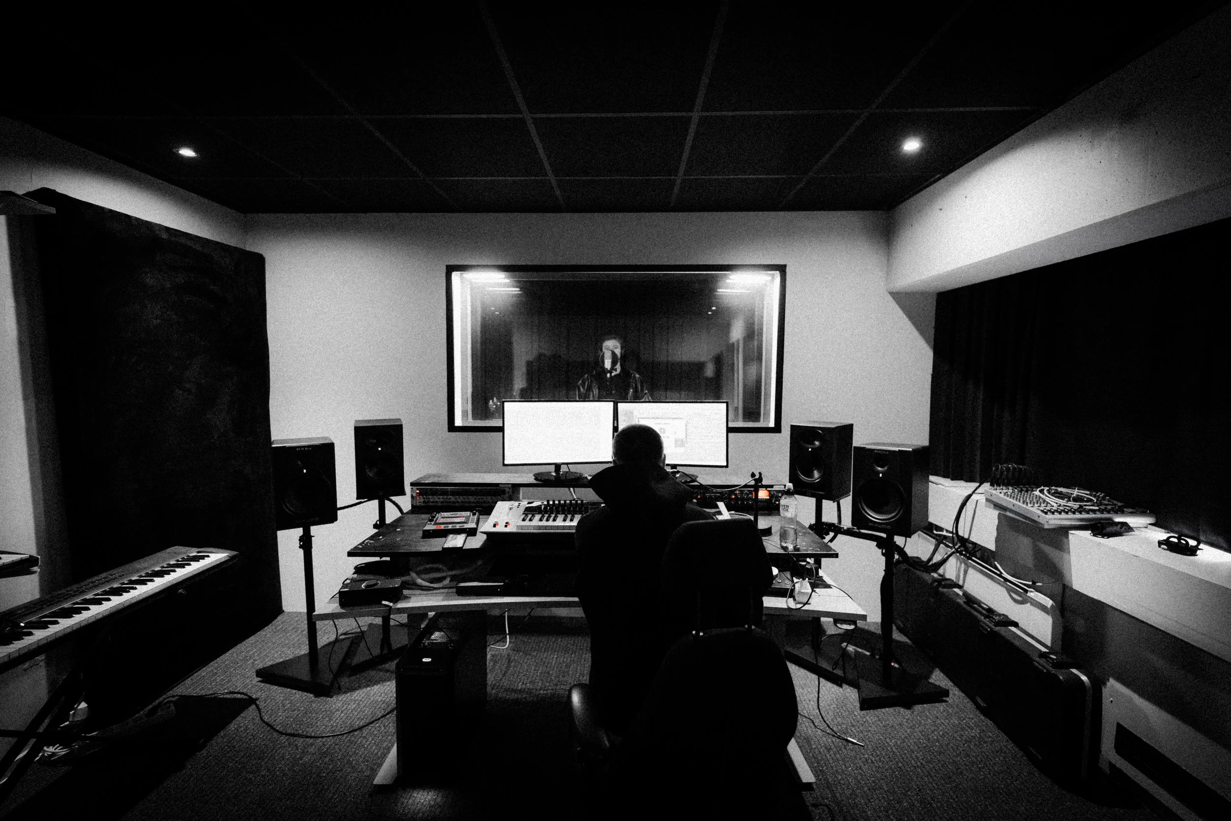 Recording studio with a person working at a mixing console in front of two monitors, a window looking into an adjacent room where a singer is being recorded.