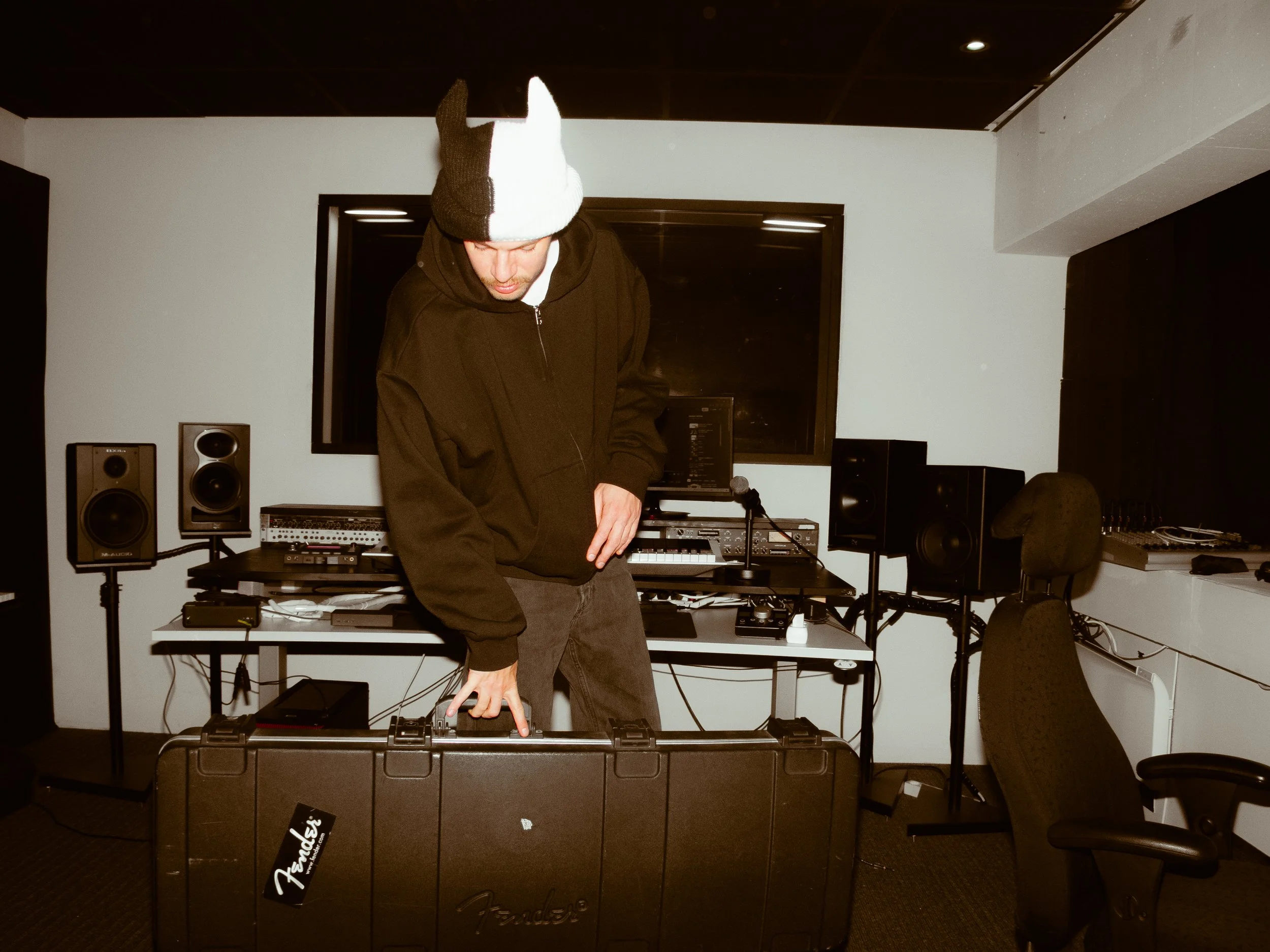 A man with a black and white knitted beanie, black hoodie, and jeans adjusting equipment inside a music studio with speakers, a computer, and audio gear.
