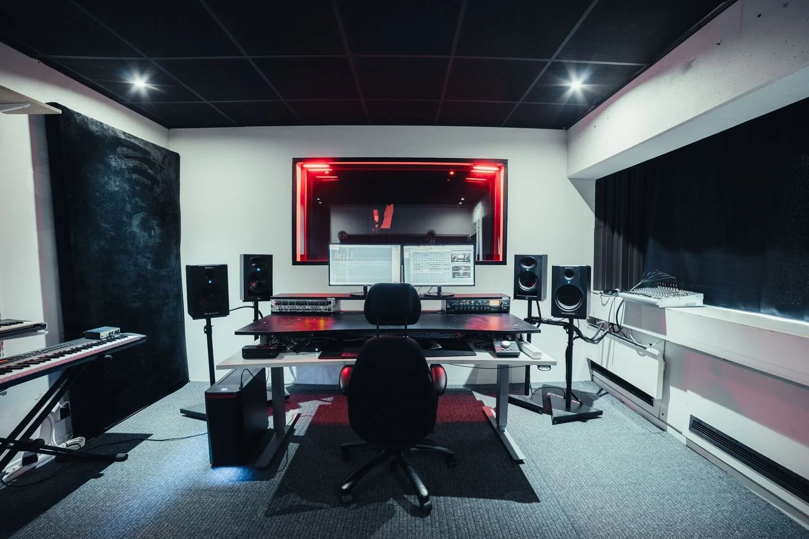 Recording studio with two computer monitors, studio speakers, keyboards, and sound equipment, illuminated by red and white lights.