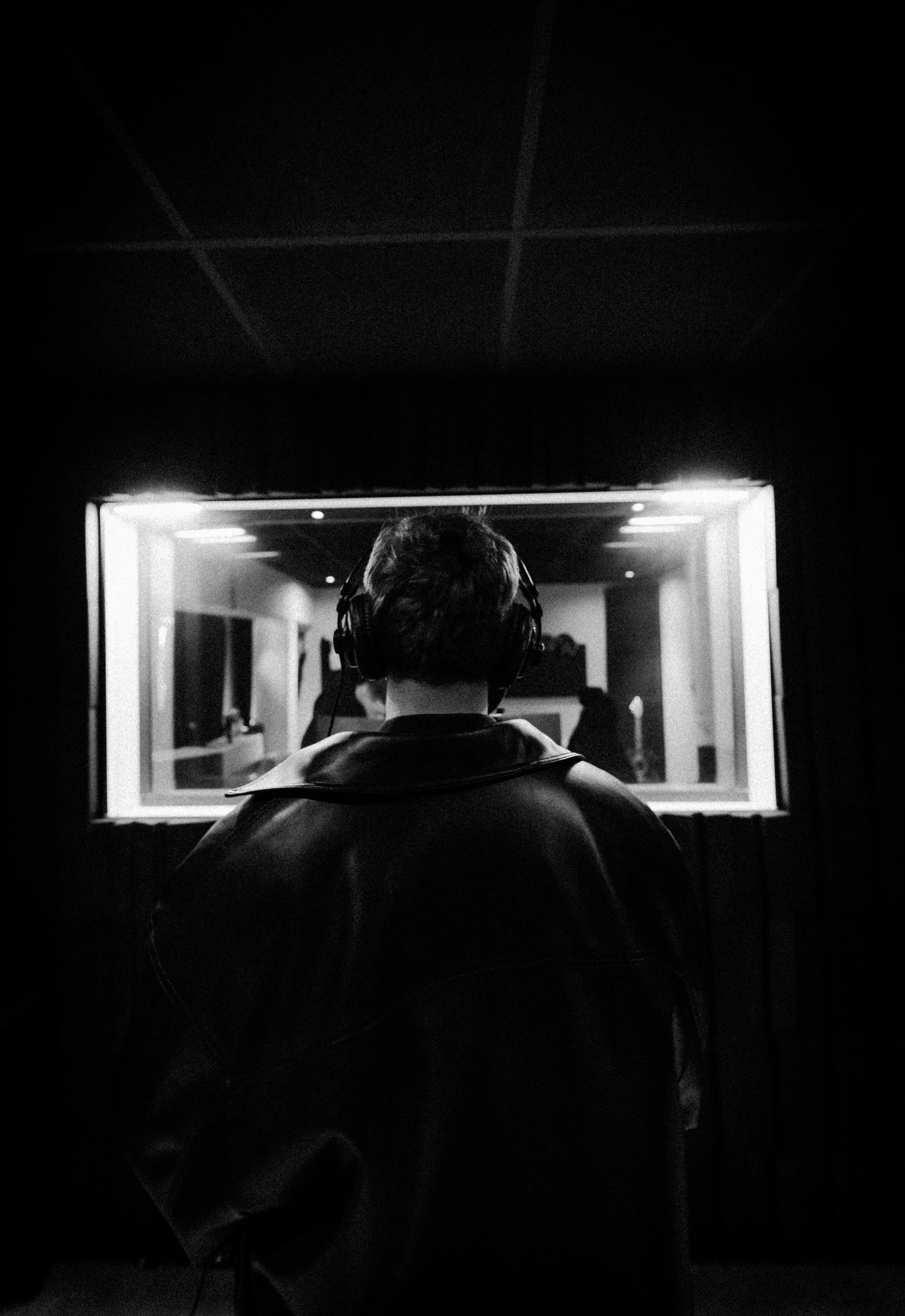 A person wearing headphones, seen from behind, in a recording studio with soundproof windows, working on audio recording.