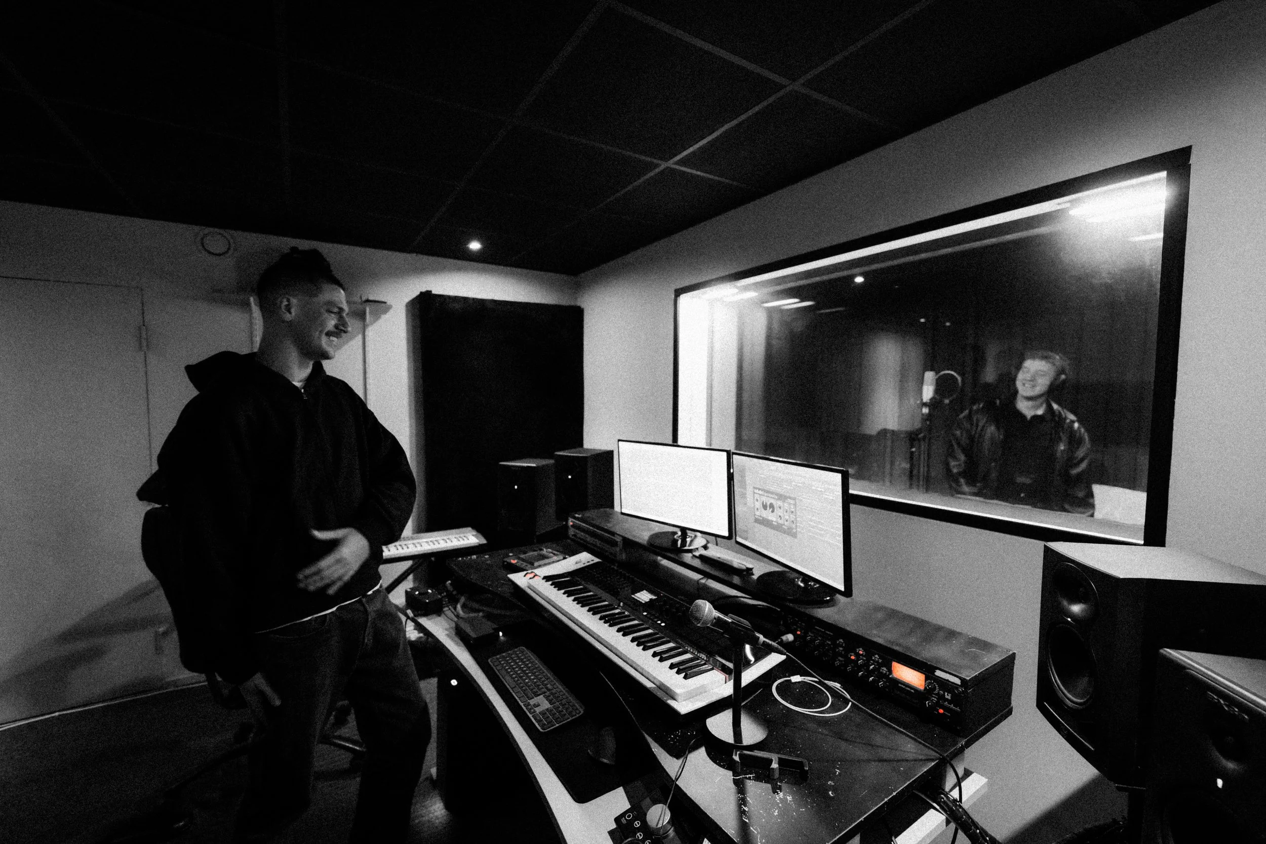 Two men in a music recording studio, one standing in front of a mixing console and the other in a vocal booth, smiling and looking at each other.