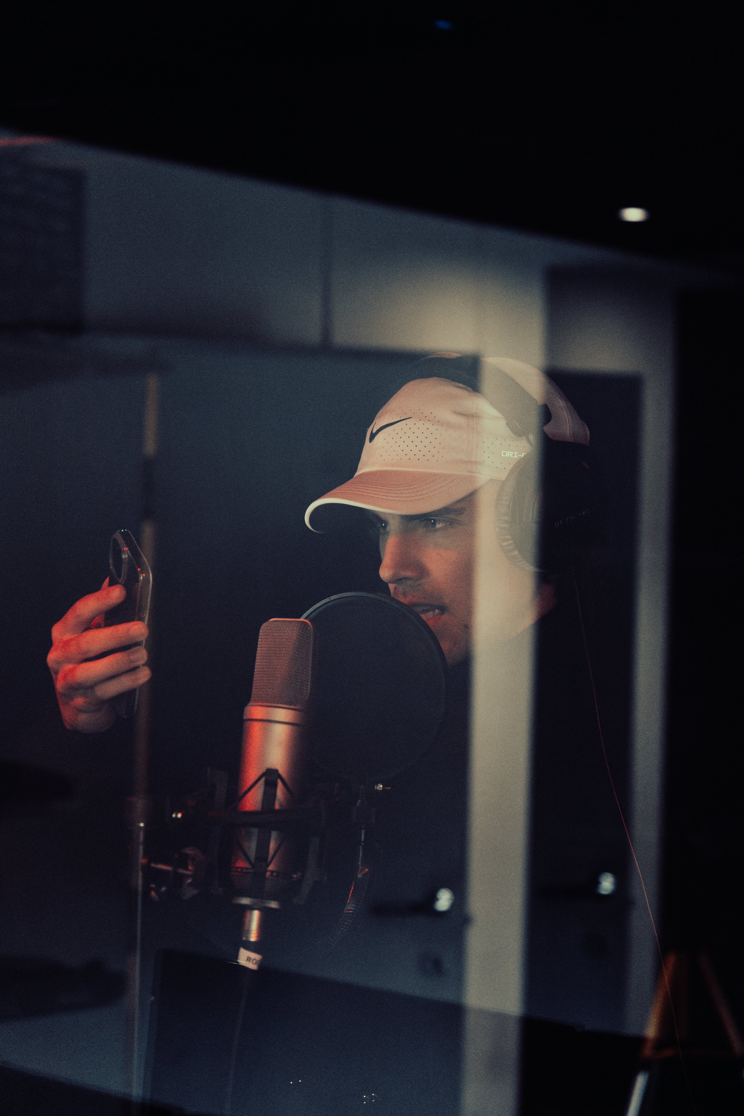 A man wearing a white Nike cap and headphones is recording vocals in a studio, seen through a glass window.