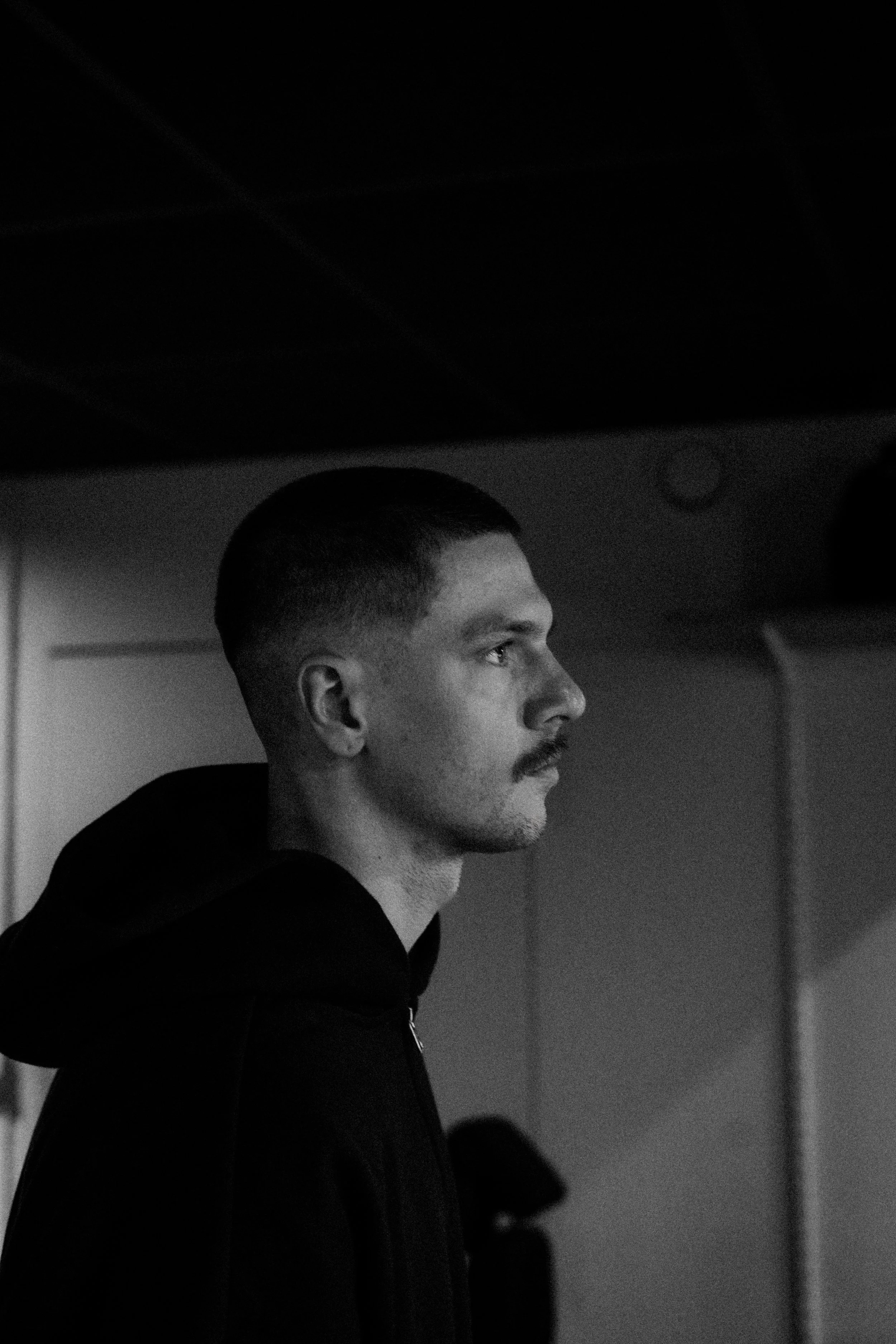 A young man with a mustache and short hair, wearing a dark hoodie, is shown in profile indoors in black and white.