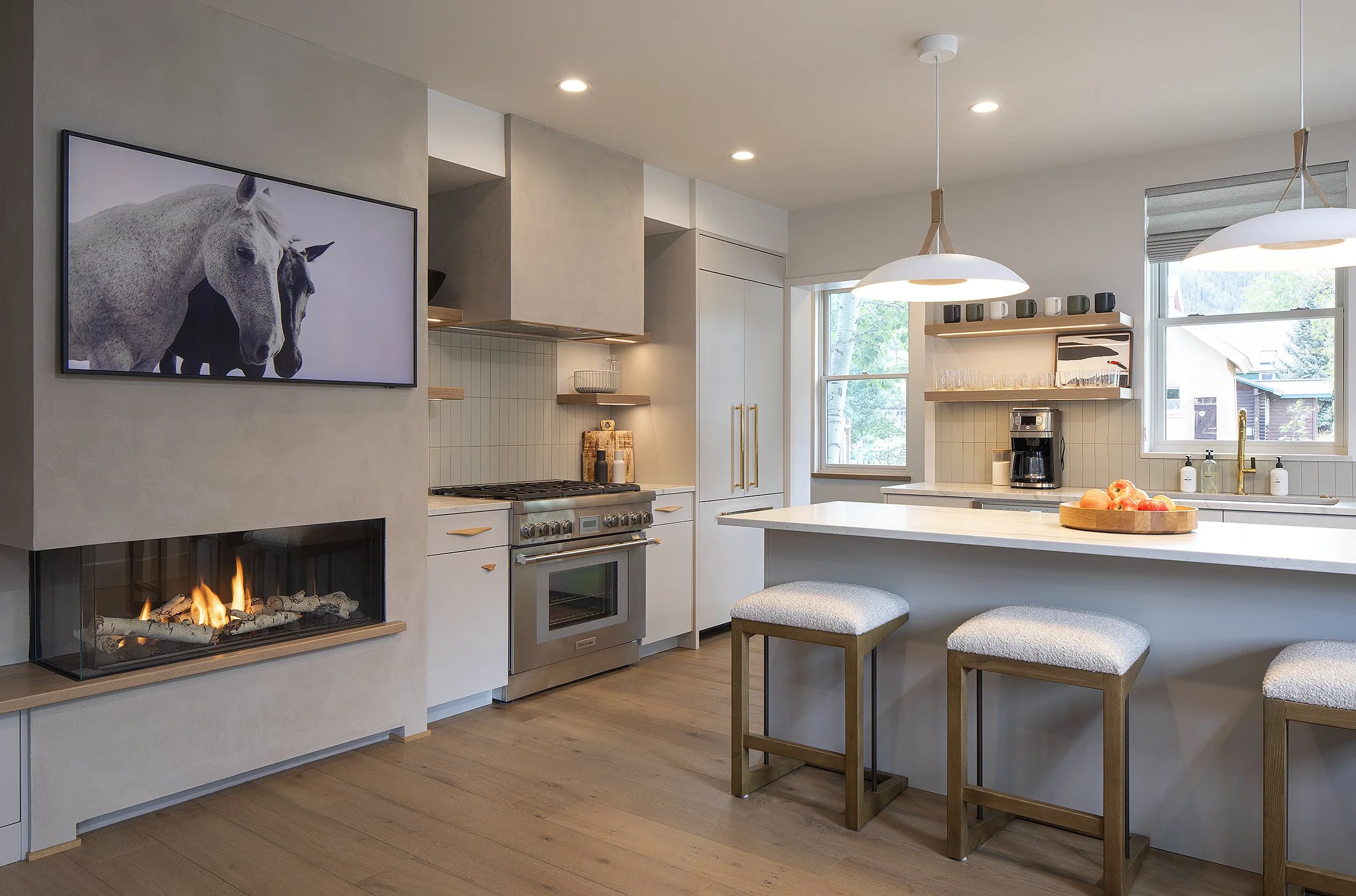 Kitchen detail with three bar stools and fruit bowel on the island with fireplace glowing and two horses on the TV monitor 