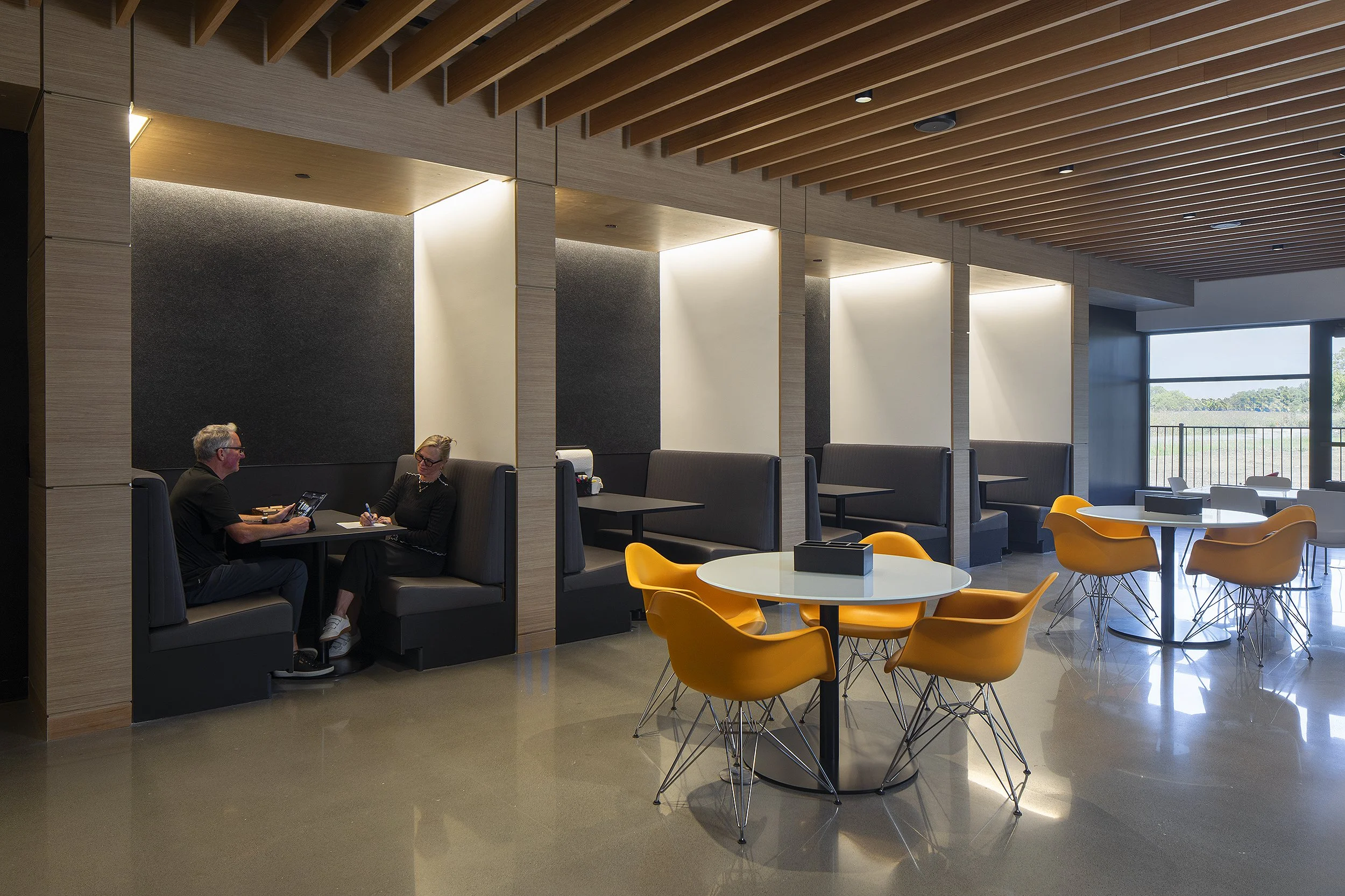 Office break room with two people sitting in a booth talking and two tables with yellow chairs