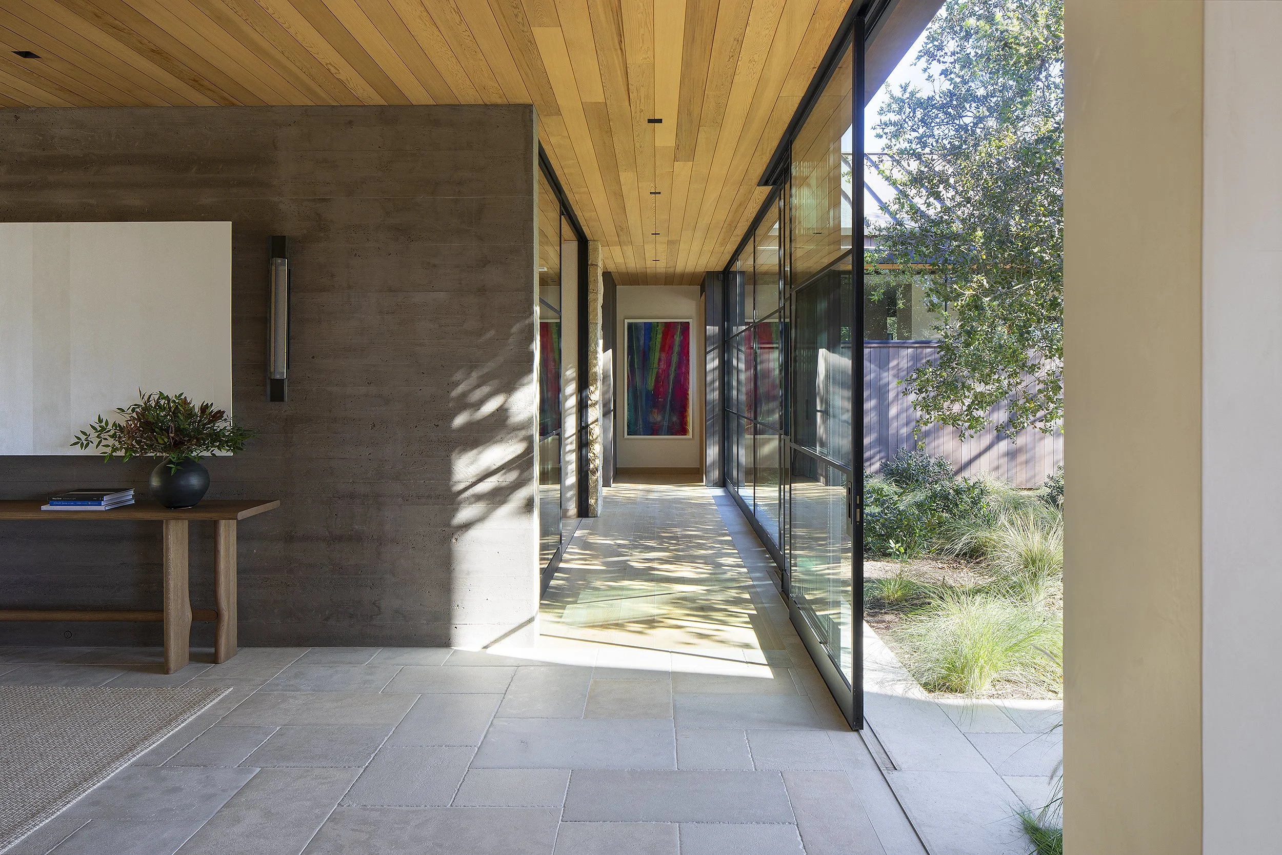 Hallway with large white painting above wooden table in entry with glass doors and walls to the right. Colorful painting a the end of the hallway Santa Barbara California 