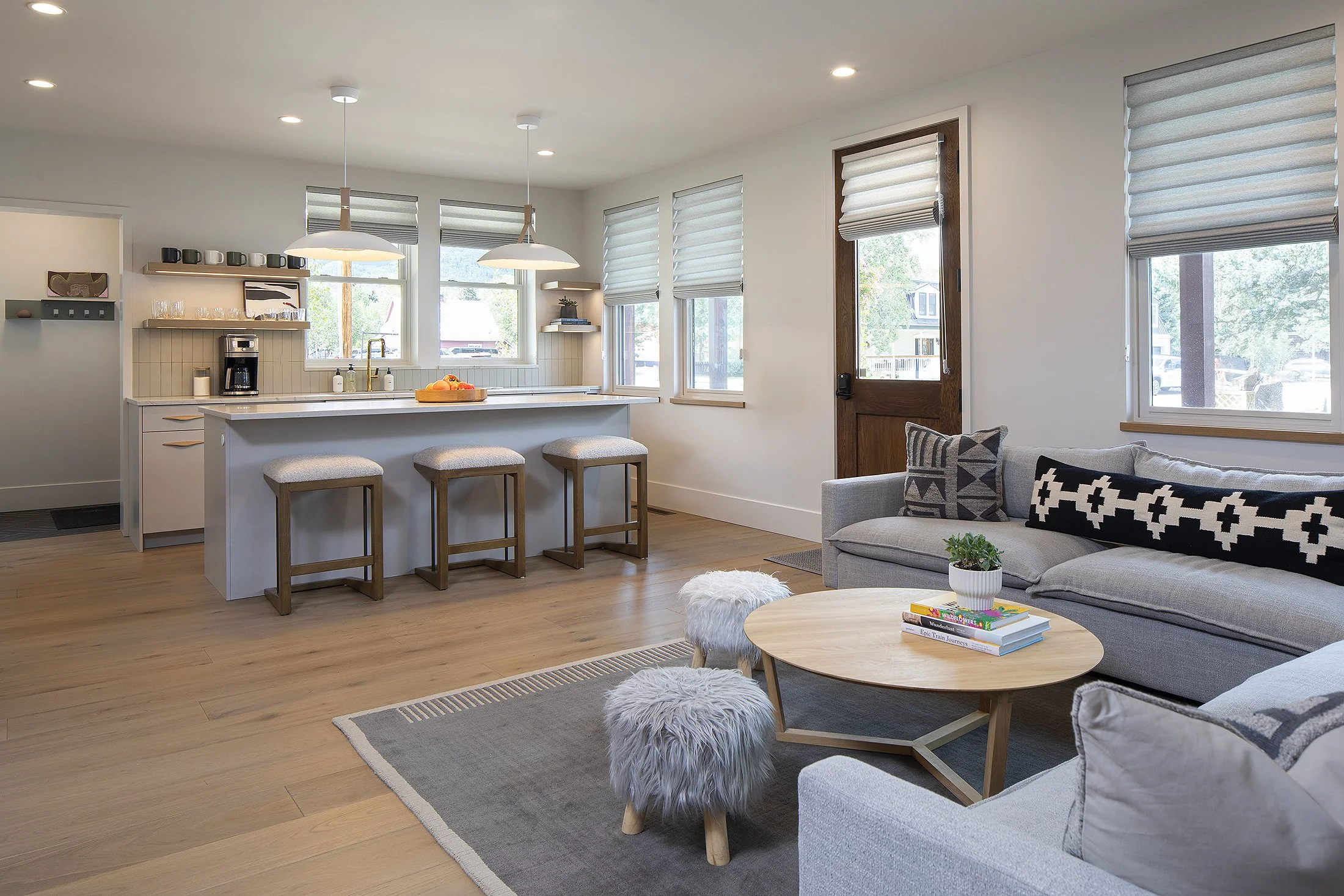 Living room with grey couches and footrests with kitchen with three bar stools at the island 