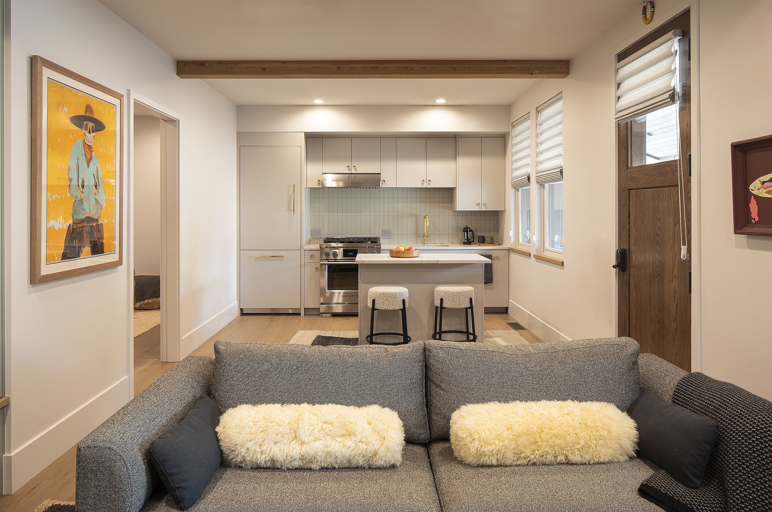 Grey couch with two while fuzzy pillows with small kitchen in the background with yellow and blue painting on wall