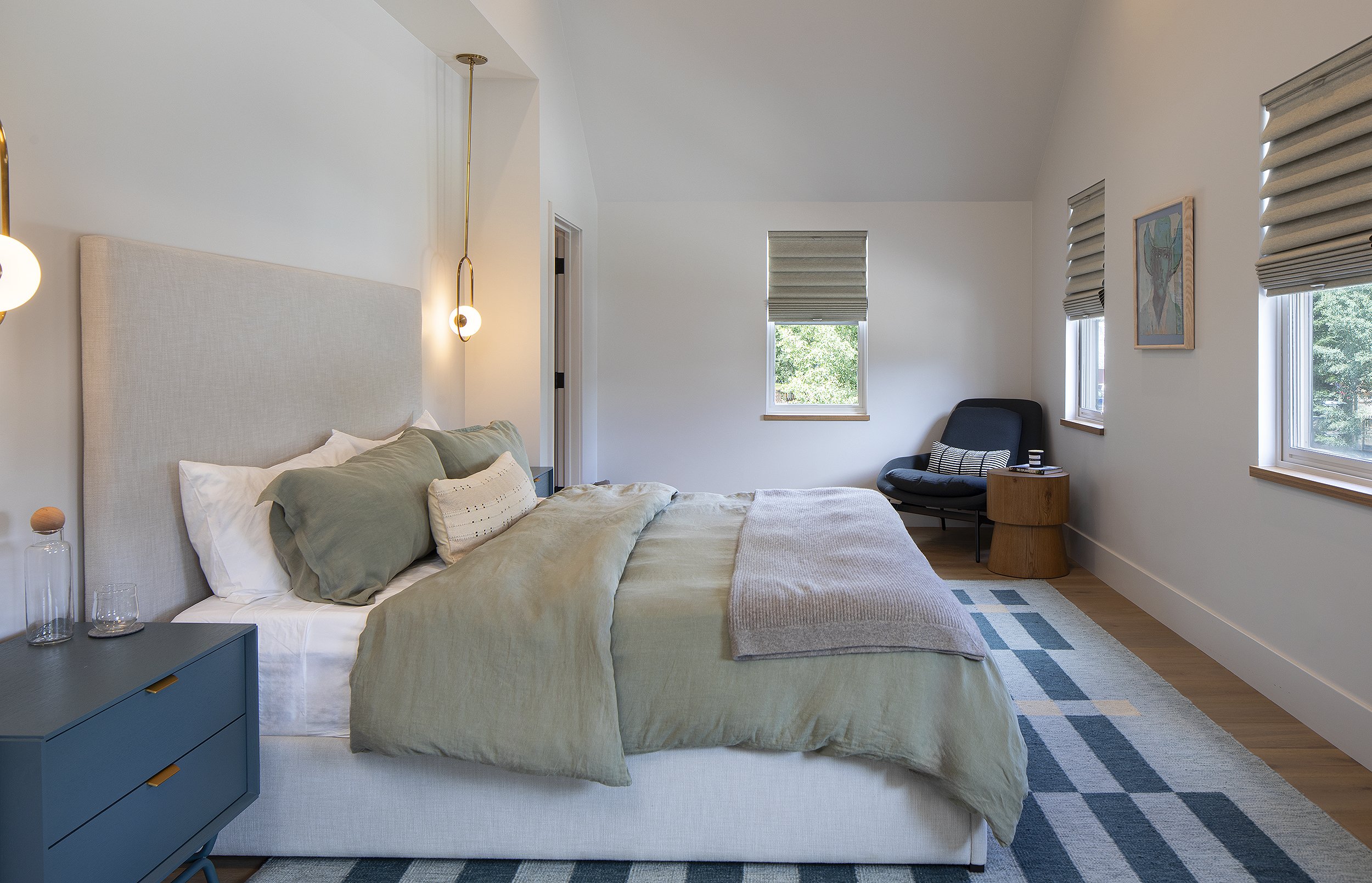 A master bedroom with  kingsize bed with green bedding and blur night stand. Chair in the corner with brown table.
