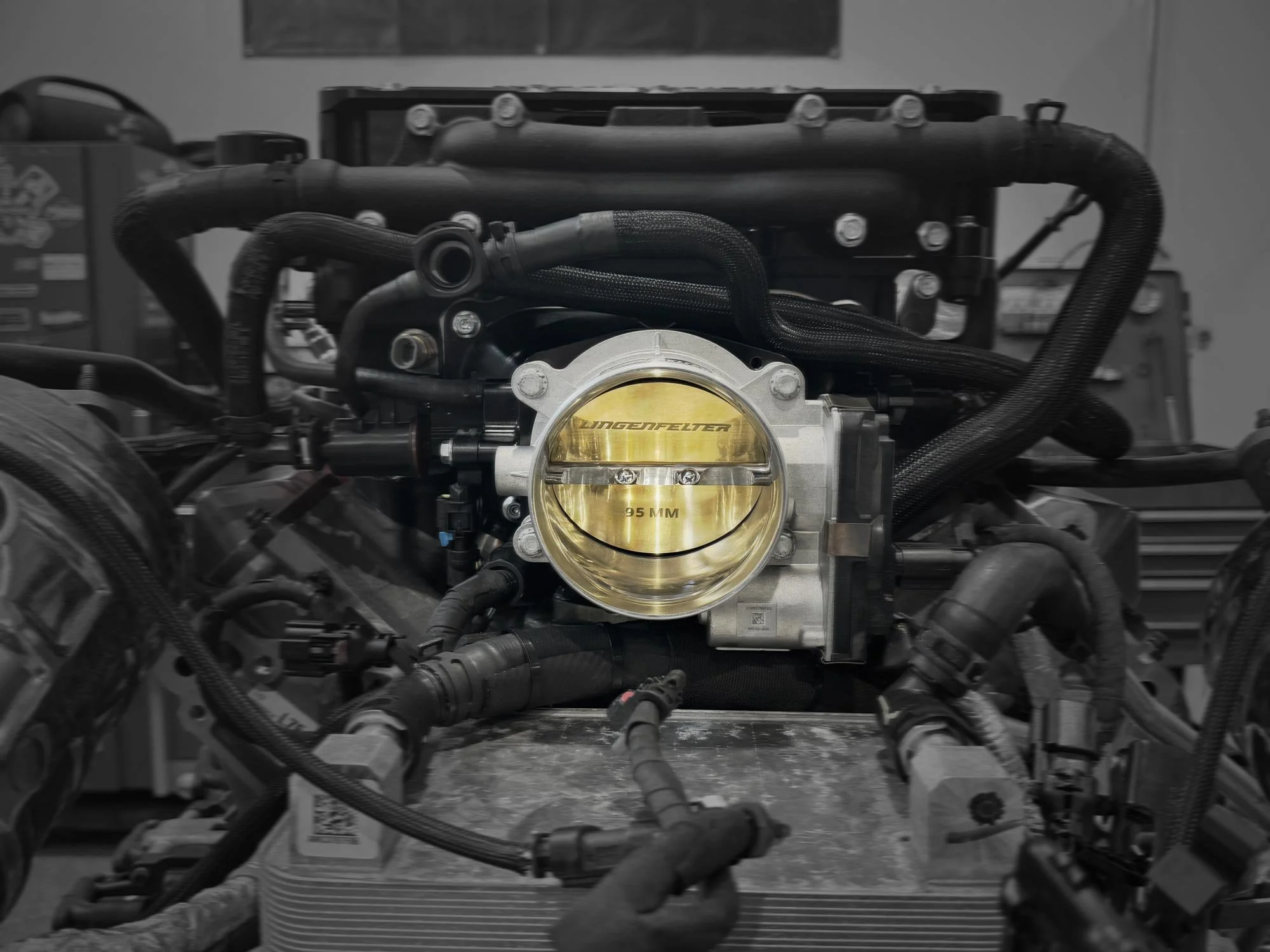 Close-up of a car engine with a yellow throttle body marked with 'Lingenfelter' and '95 MM', surrounded by black hoses and electrical wiring.