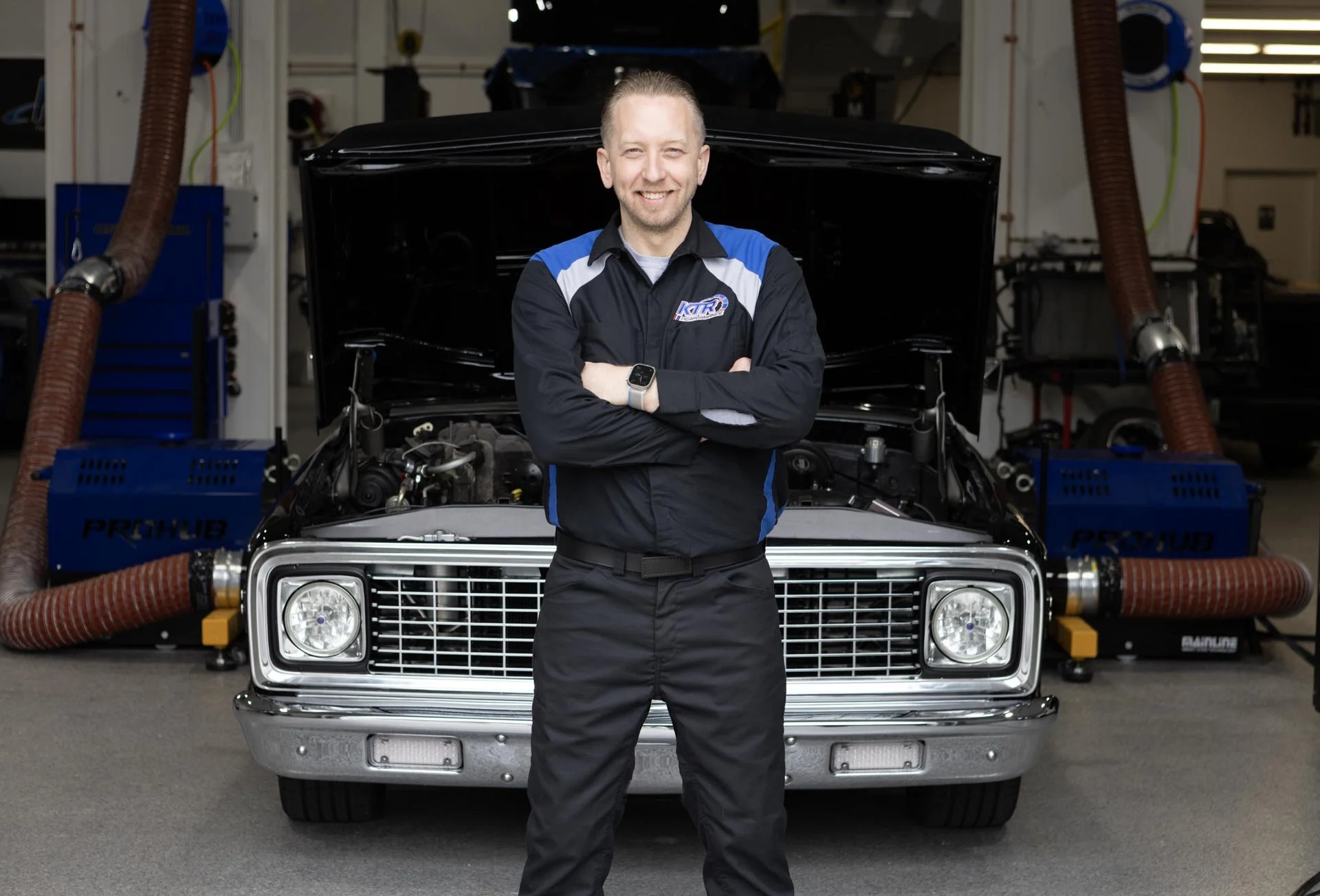 Master Mechanic and Owner of KTR Perforance, Kyle, in a black and blue shop uniform standing with arms crossed in front of a classic 1972 Chevrolet C10 with an open hood inside a garage or workshop.