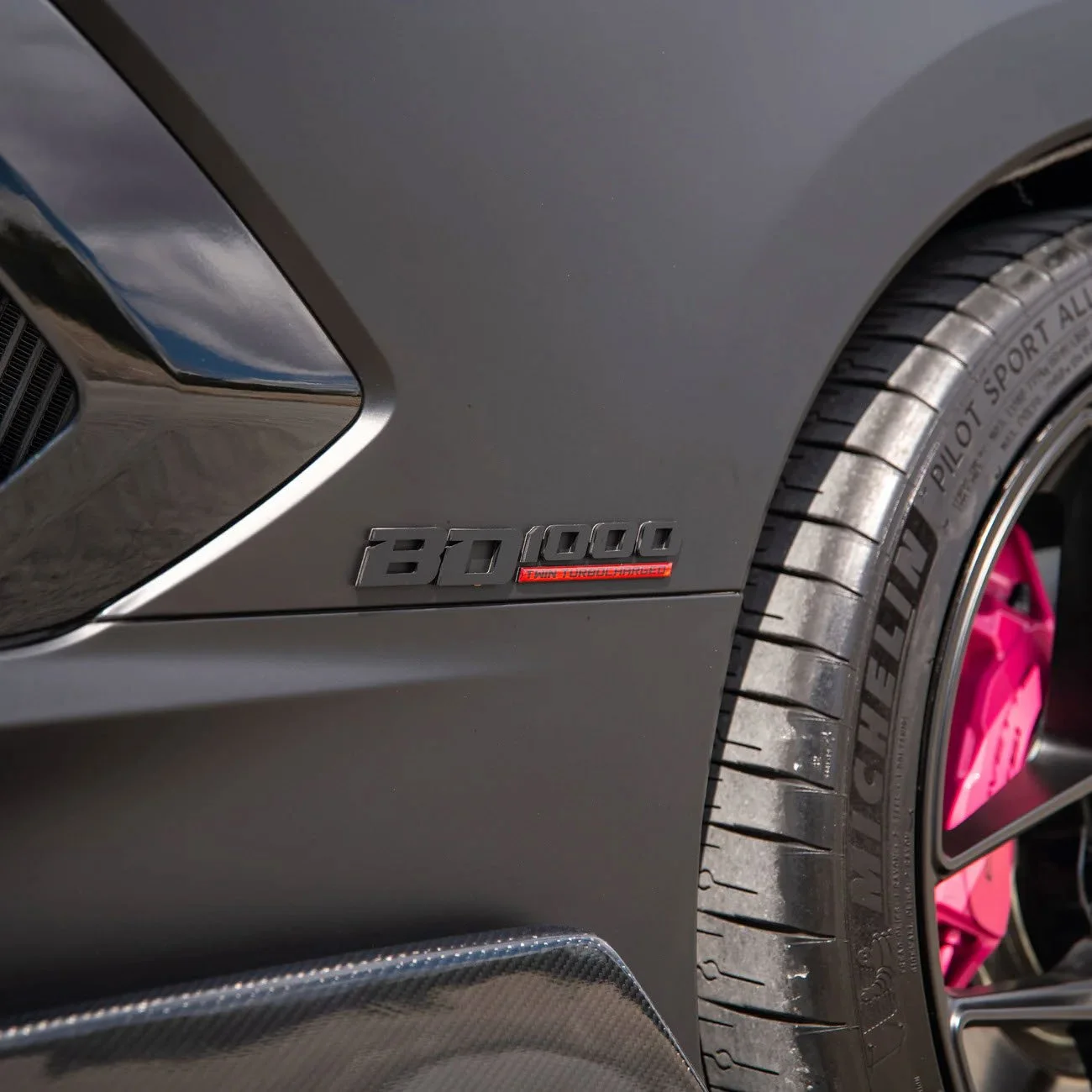 Close-up of a grey sports car with a 'B0 1000' badge and red 'Twin Turbo V8' badge, showing part of the front bumper, tire, and pink wheel rim.