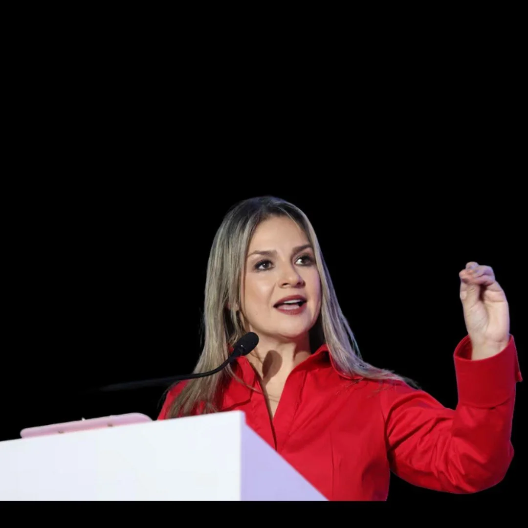 Una mujer hablando en un micrófono, con expresión decidida, vestida con una camisa roja, frente a un fondo negro.