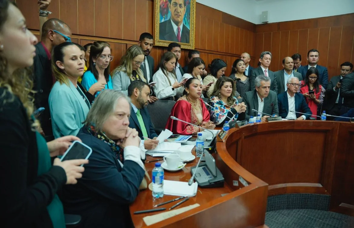 Varias personas en una sala de reuniones, algunas sentadas en una mesa con micrófonos y agua, otras de pie a su alrededor, en un entorno formal con retrato de un hombre en la pared.