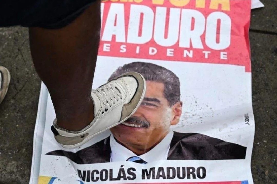 Persona de pie sobre un periódico que muestra la imagen del expresidente Nicolás Maduro y el nombre "Nicolás Maduro" en la parte inferior.