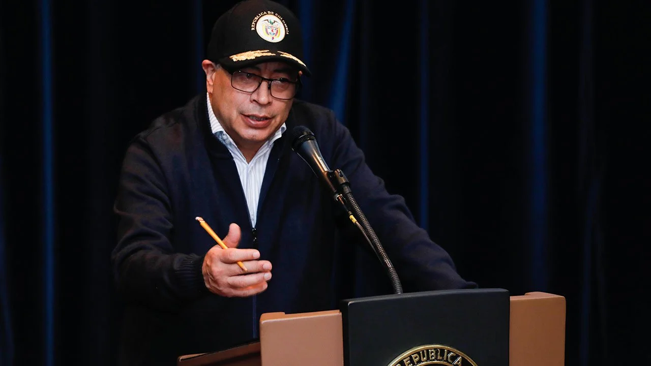 hombre hablando en un podio con micrófono, usa gafas, gorra con escudo y ropa oscura, fondo negro