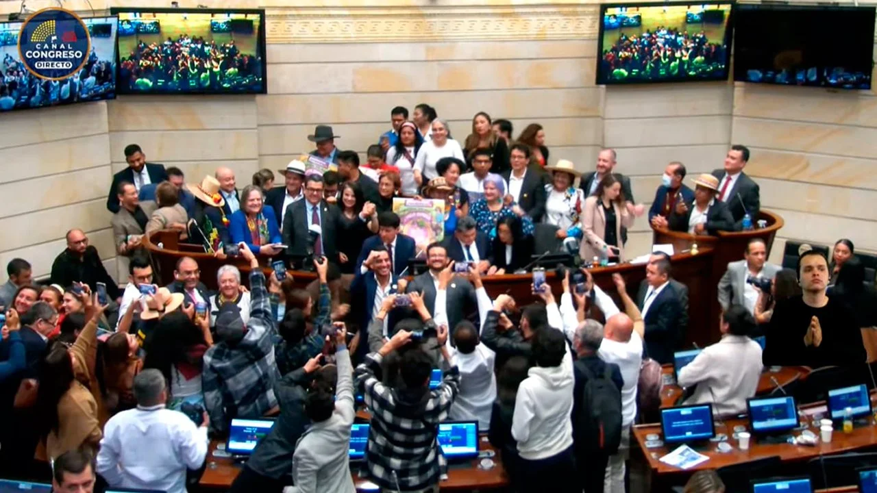 Reunión en una sala del Congreso con muchas personas, algunos toman fotos y otros hacen aplausos, en la pantalla aparecen varias imágenes de una multitud.