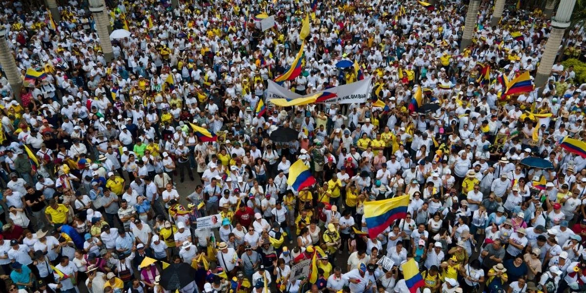 Gran multitud en una manifestación con banderas de Colombia, sosteniendo pancartas y paraguas.