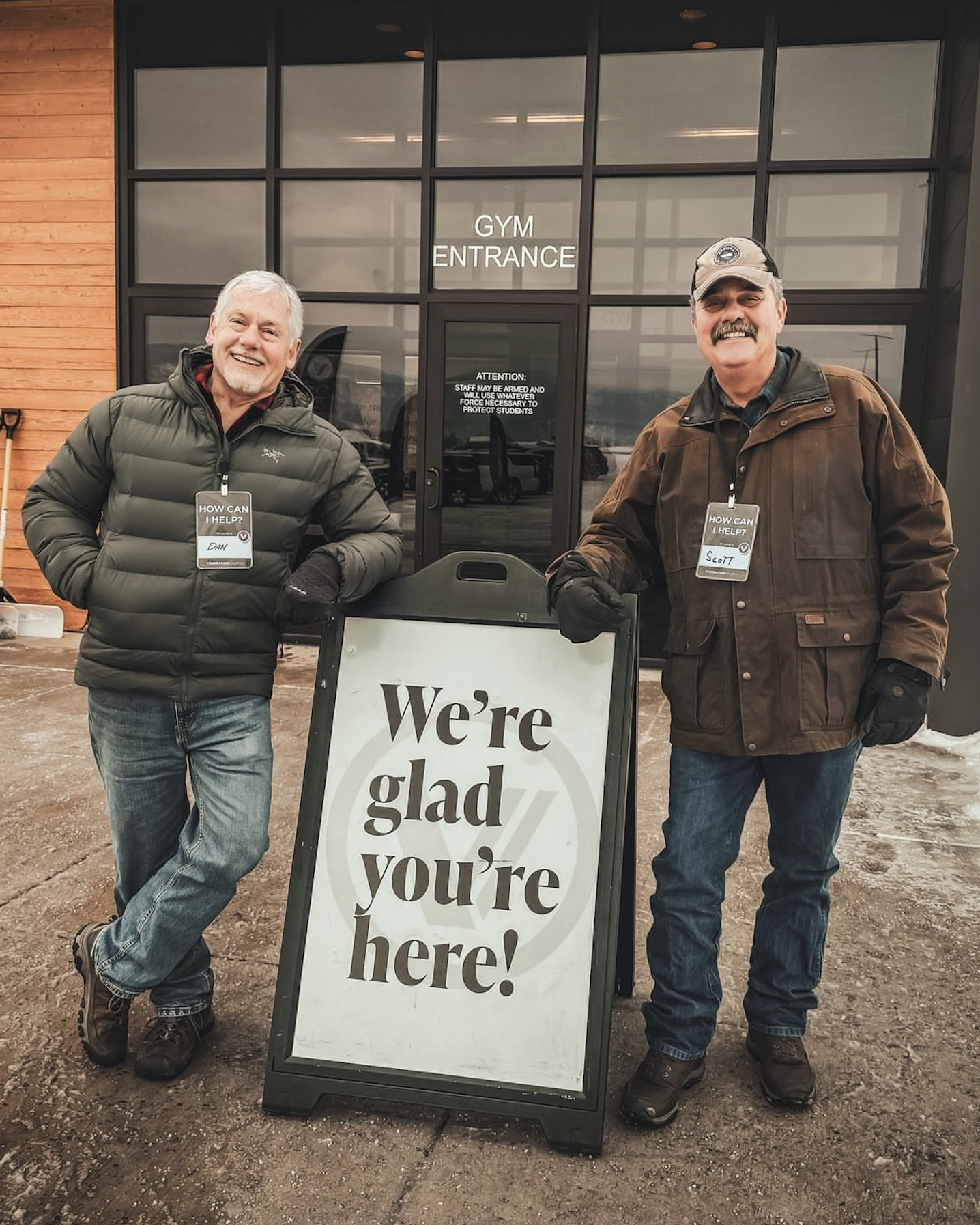 Two smiling men wearing jackets and name tags standing next to a sign that says 'We're glad you're here!' Outside a gym entrance.