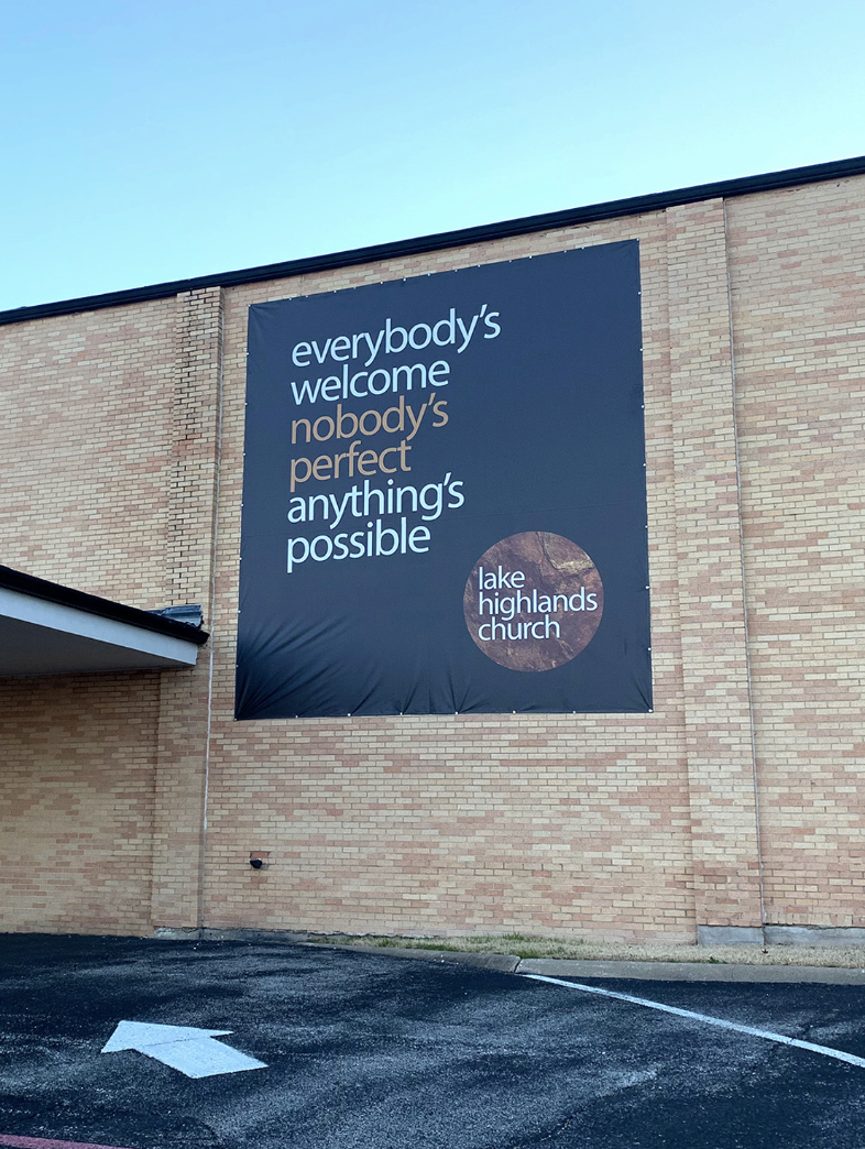 Large black banner on a brick building exterior displaying the message 'everybody's welcome, nobody's perfect, anything's possible' along with the church name 'lake highlands church' in a circular logo.