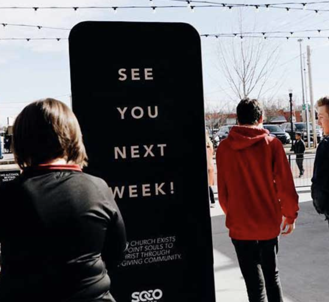A black sign with white text saying "See You Next Week!" is displayed outdoors, with people walking past on a sidewalk.