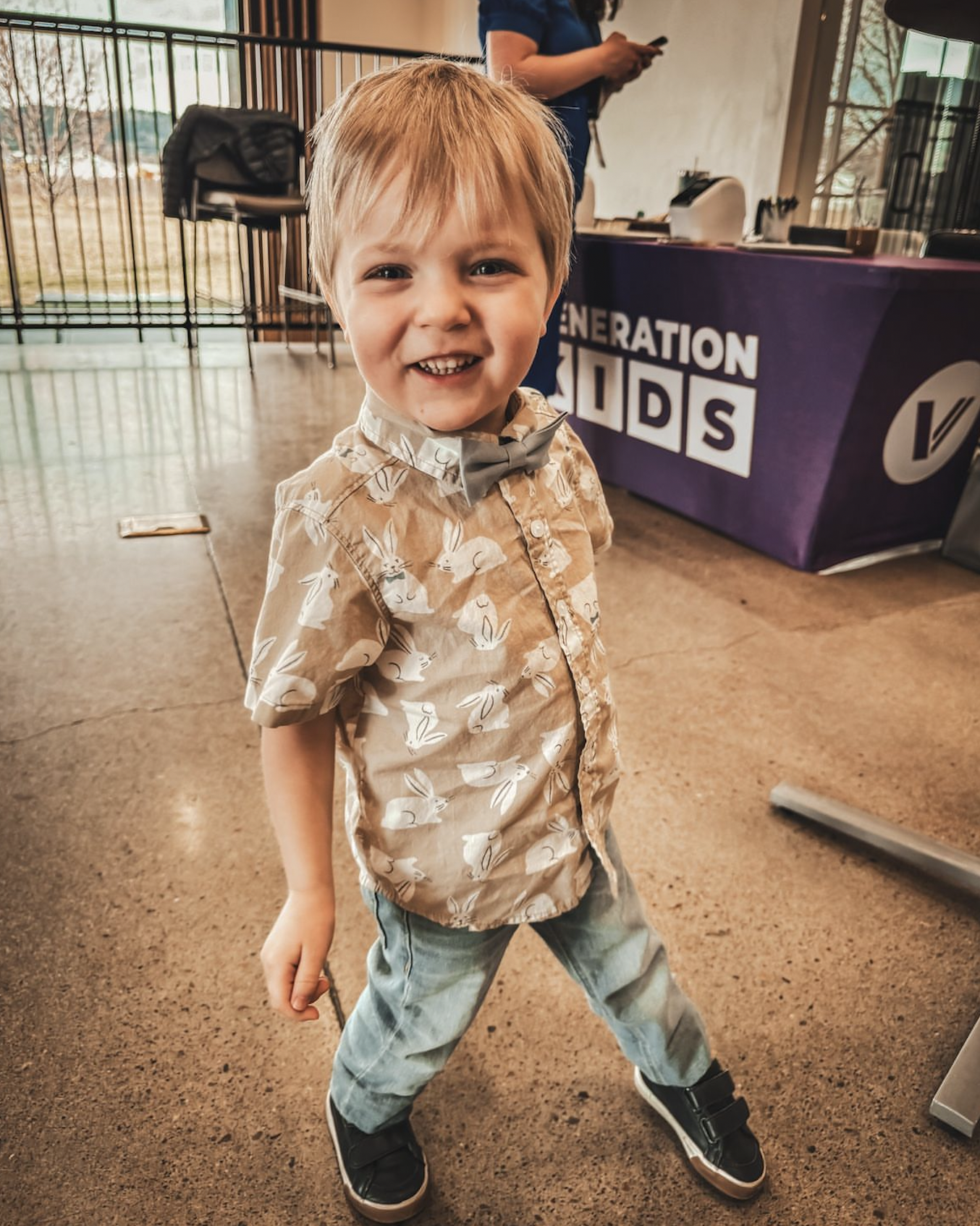 Smiling young boy with light brown hair, wearing a tan shirt with white bunny patterns, gray bow tie, distressed jeans, and black sneakers, standing indoors in front of a purple table with banners and a woman in the background.
