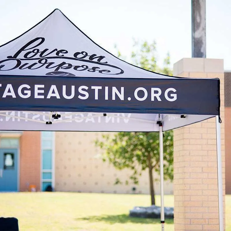 A white and black canopy tent with the website 'AGEAUSTIN.ORG' on it, outdoors on a sunny day, with a tree and building in the background.