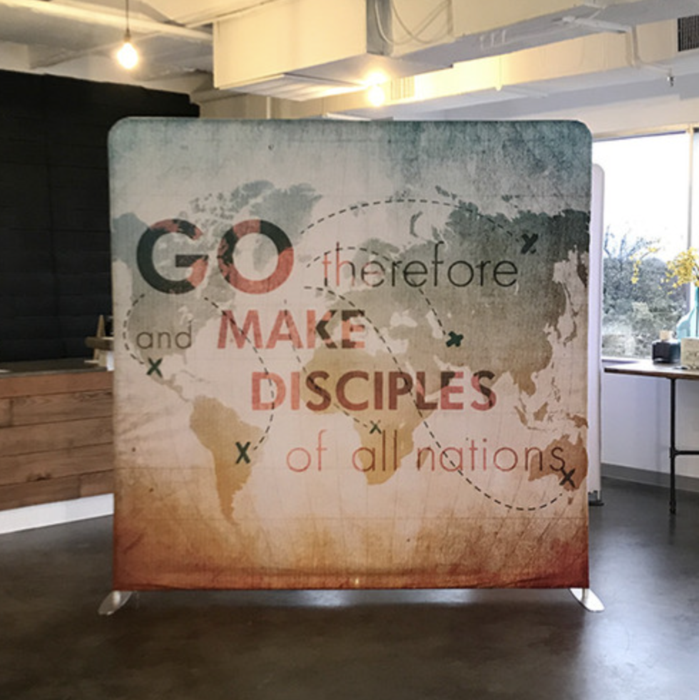A world map with the quote "Go therefore and make disciples of all nations" written over it, displayed on a stand in an indoor space with windows on the right.