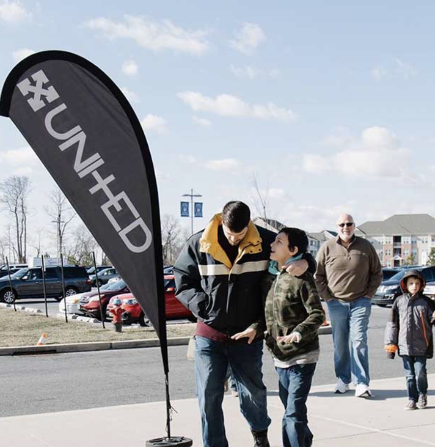 A group of people walking outdoors on a sunny day near parked cars, with a large black flag that reads 'UNHED' in the foreground.