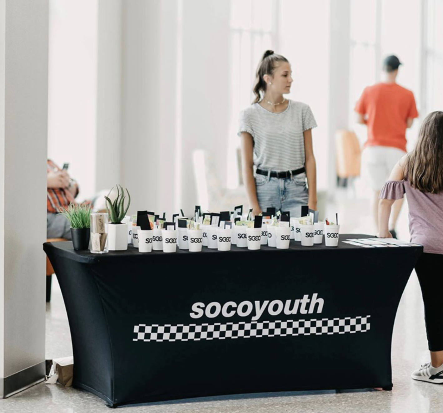 A woman standing behind a table with 'socoyouth' printed on it, with cups and small plant pots on display, at an indoor event with other people in the background.