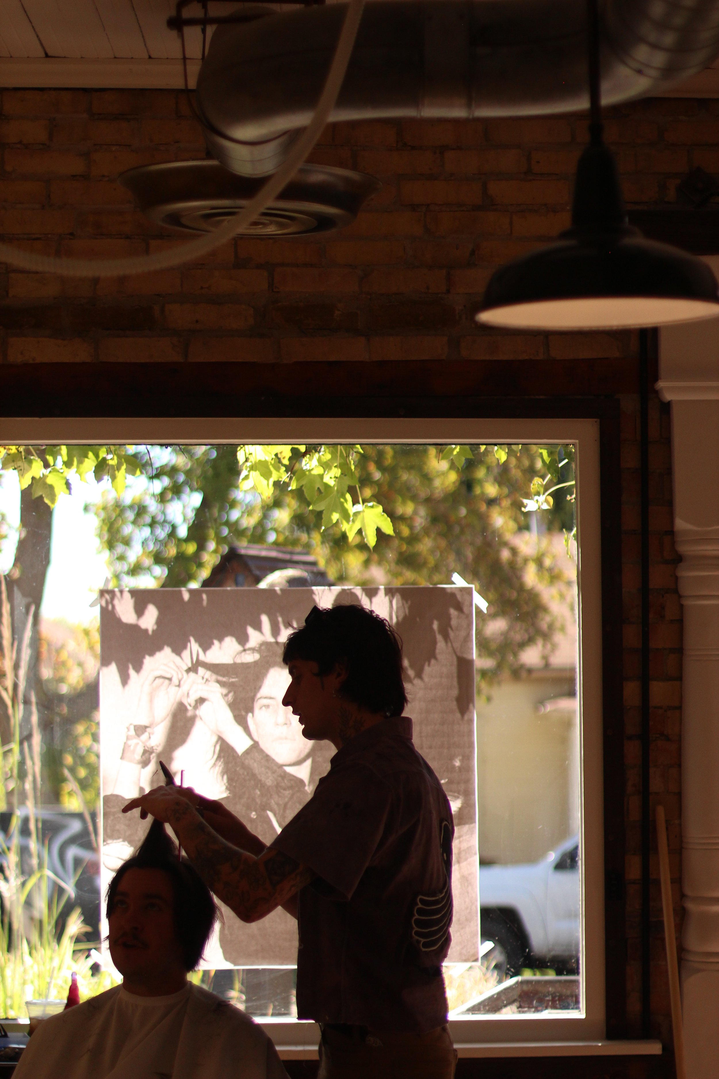 A hairstylist working on a client's hair inside a salon, with a large window showing trees and sunlight outside and photo of patti smith