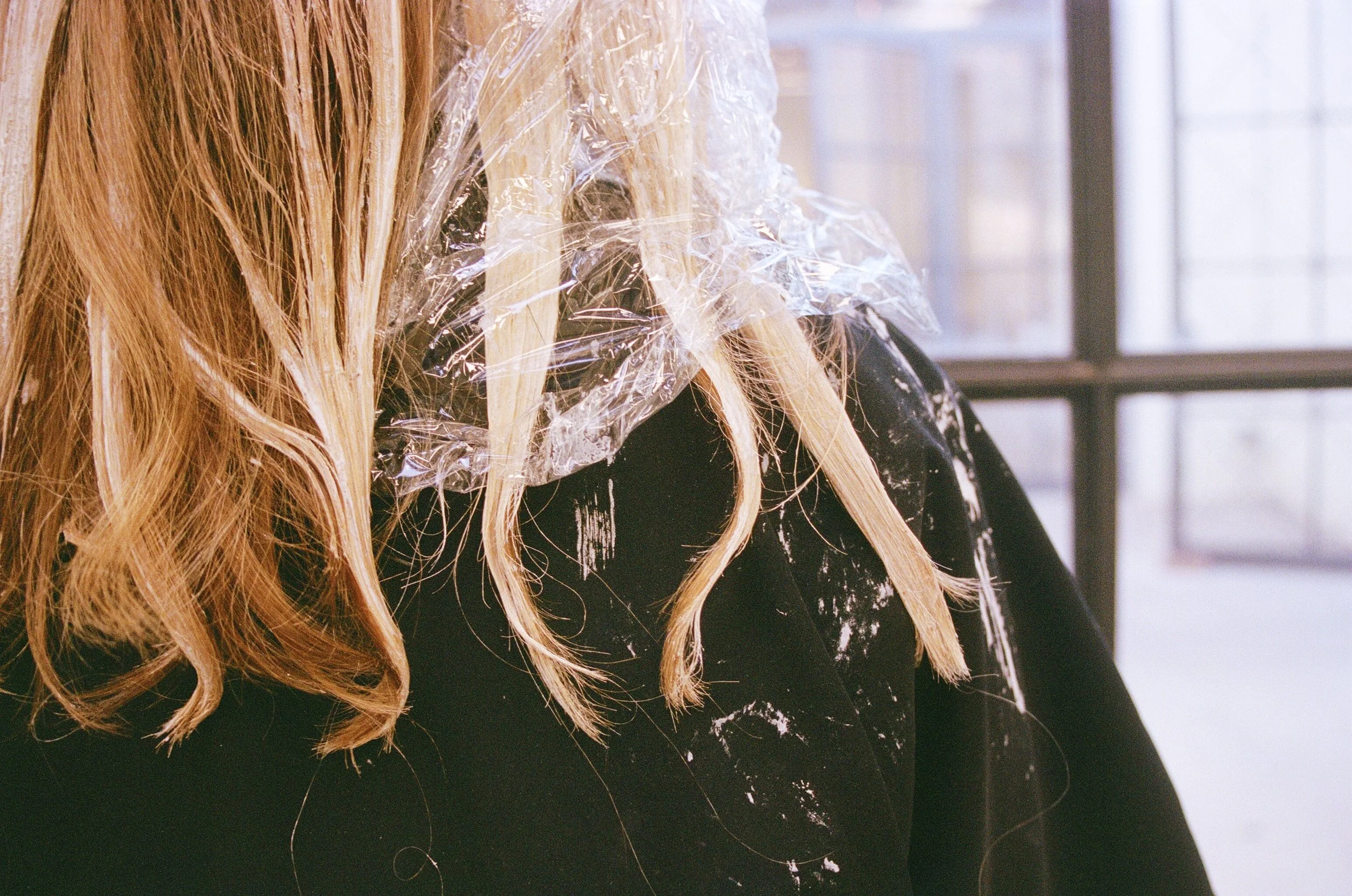 Close-up of a person with long blonde hair wearing a black shirt, with some hair and shirt covered in white paint splatters, and a clear plastic sheet partially covering their hair.