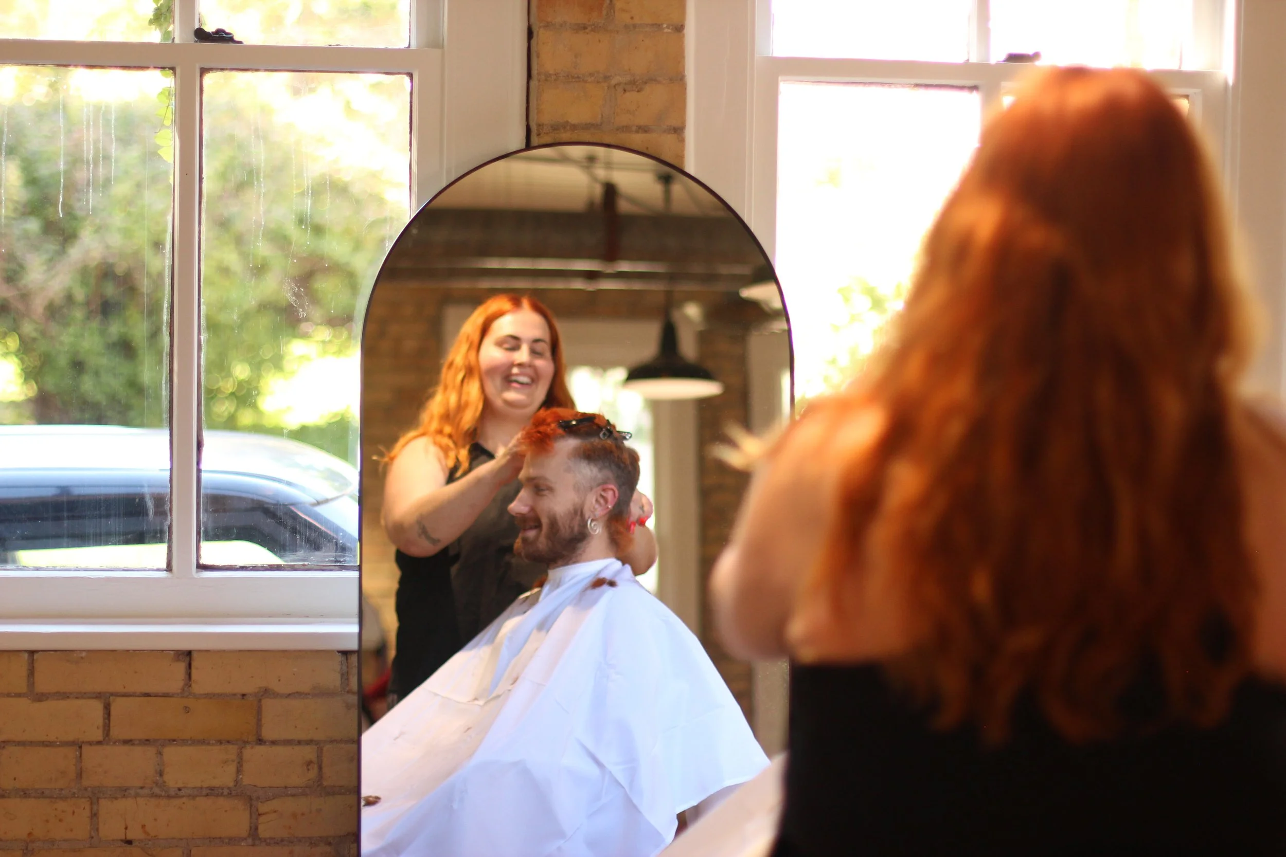 A woman with red hair is giving a haircut to a man with short hair and a beard in a salon. They are both smiling, and the scene is reflected in a large mirror.