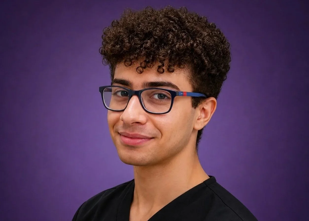 A young man with curly dark hair, glasses, and a slight smile, wearing a black shirt, against a purple background.