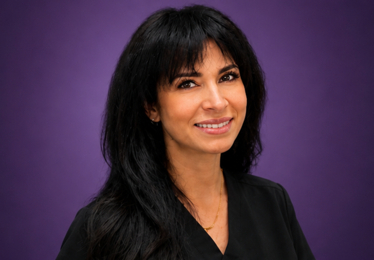 Portrait of a woman with long black hair, wearing a black top and a gold necklace, smiling against a purple background.