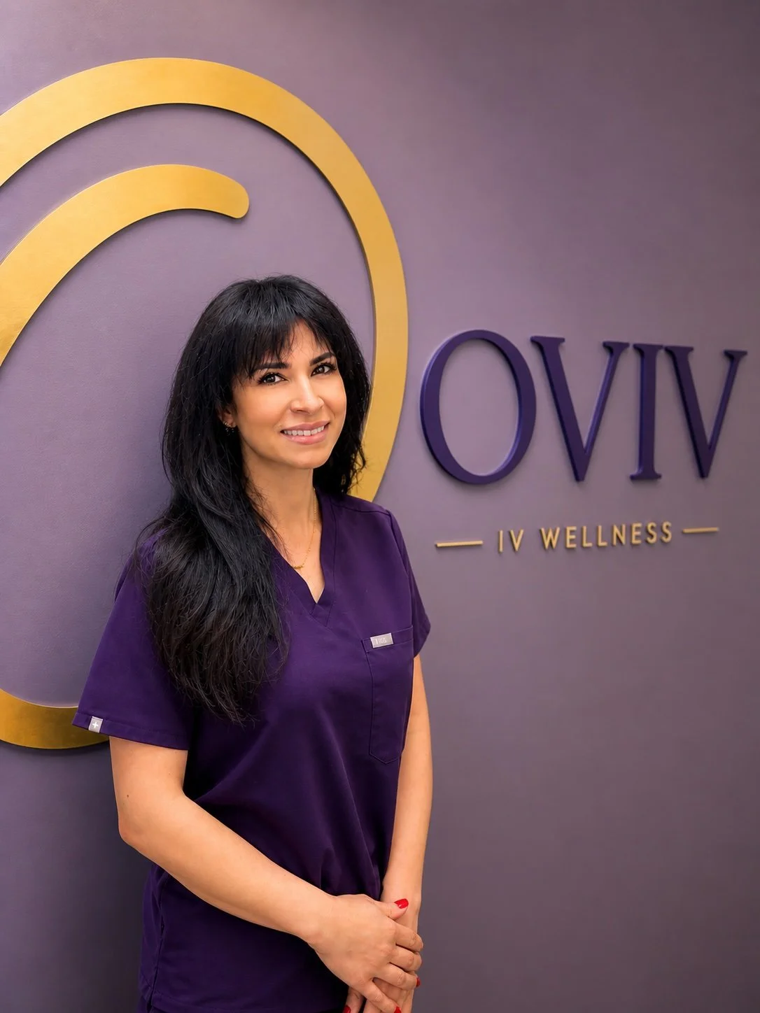 A woman with long black hair wearing purple scrubs standing in front of a wall with the logo and name 'OVIV IV WELLNESS' in purple and gold.