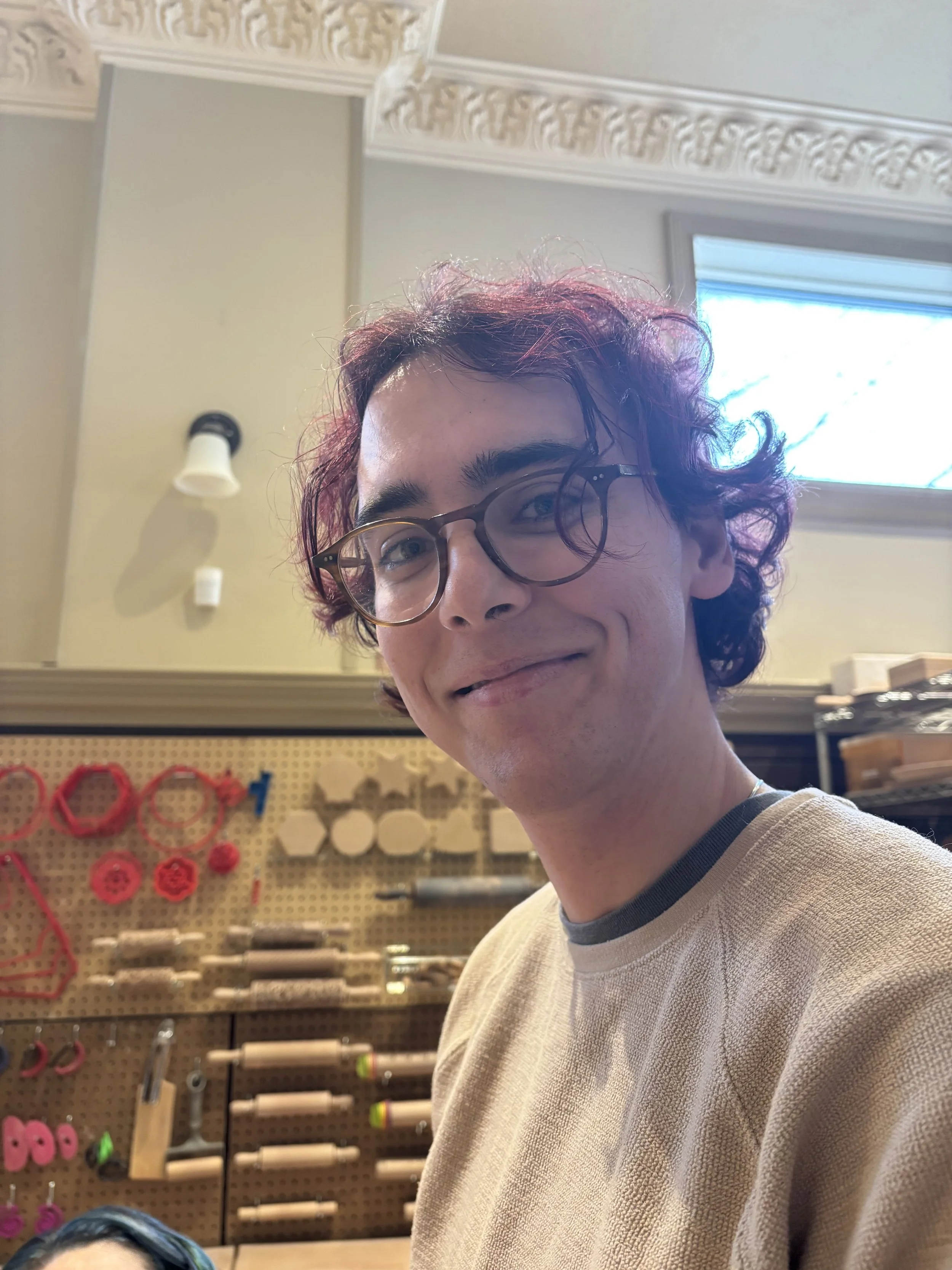 E. Enbom (they/them) is a white person with dyed red hair and glasses. They are smiling in front of a row of pottery tools.