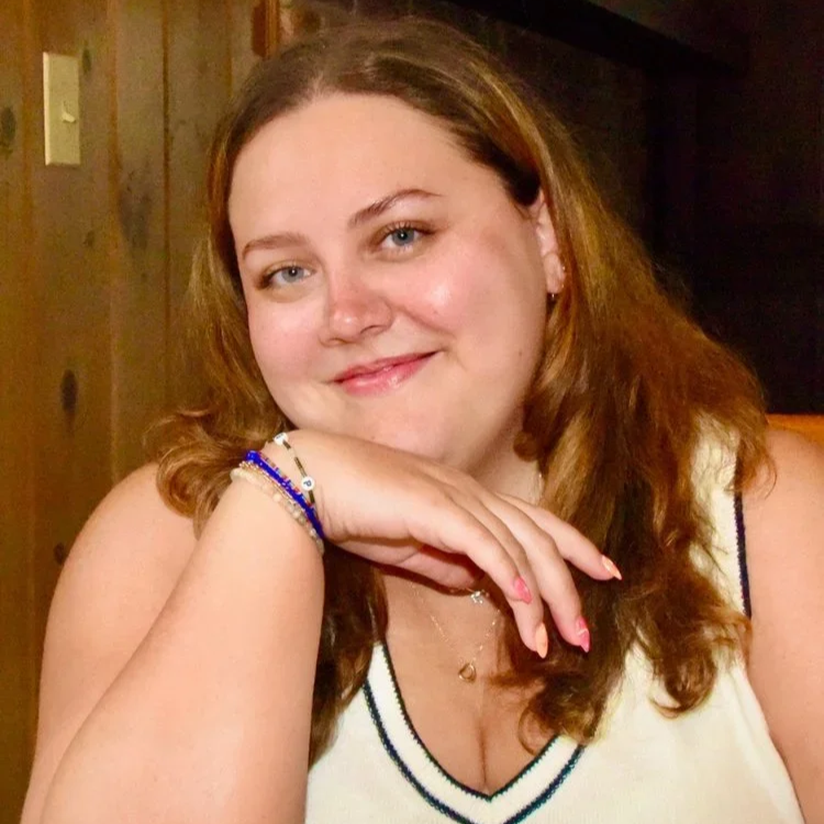 Lexi Rosen (she/her) is a white, Greek American woman with long, curly brown hair and blue eyes. She is smiling.