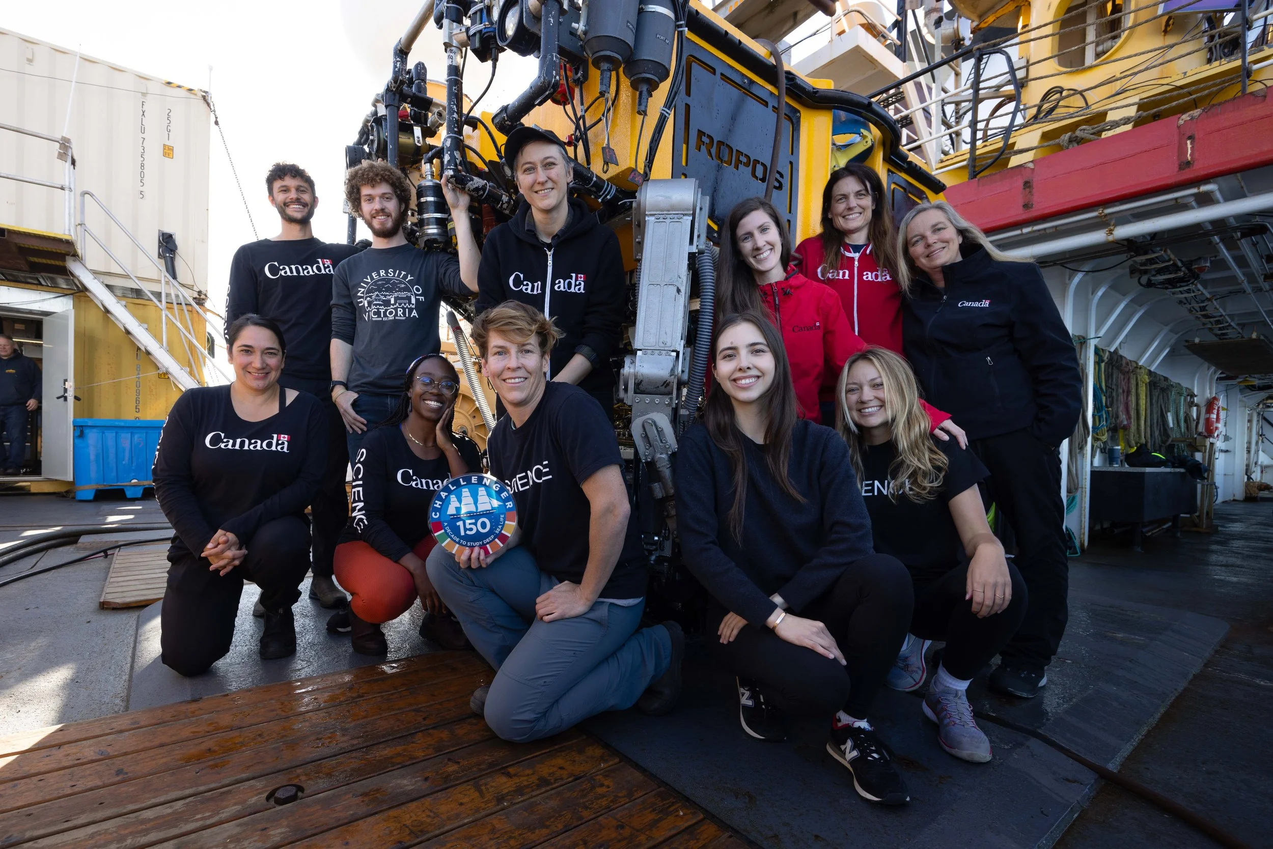 The at sea team is gathered around the remotely operated vehicle (ROV) ROPOS.  Team lead Cherisse Du Preez is holding a Challenger 150 sign.