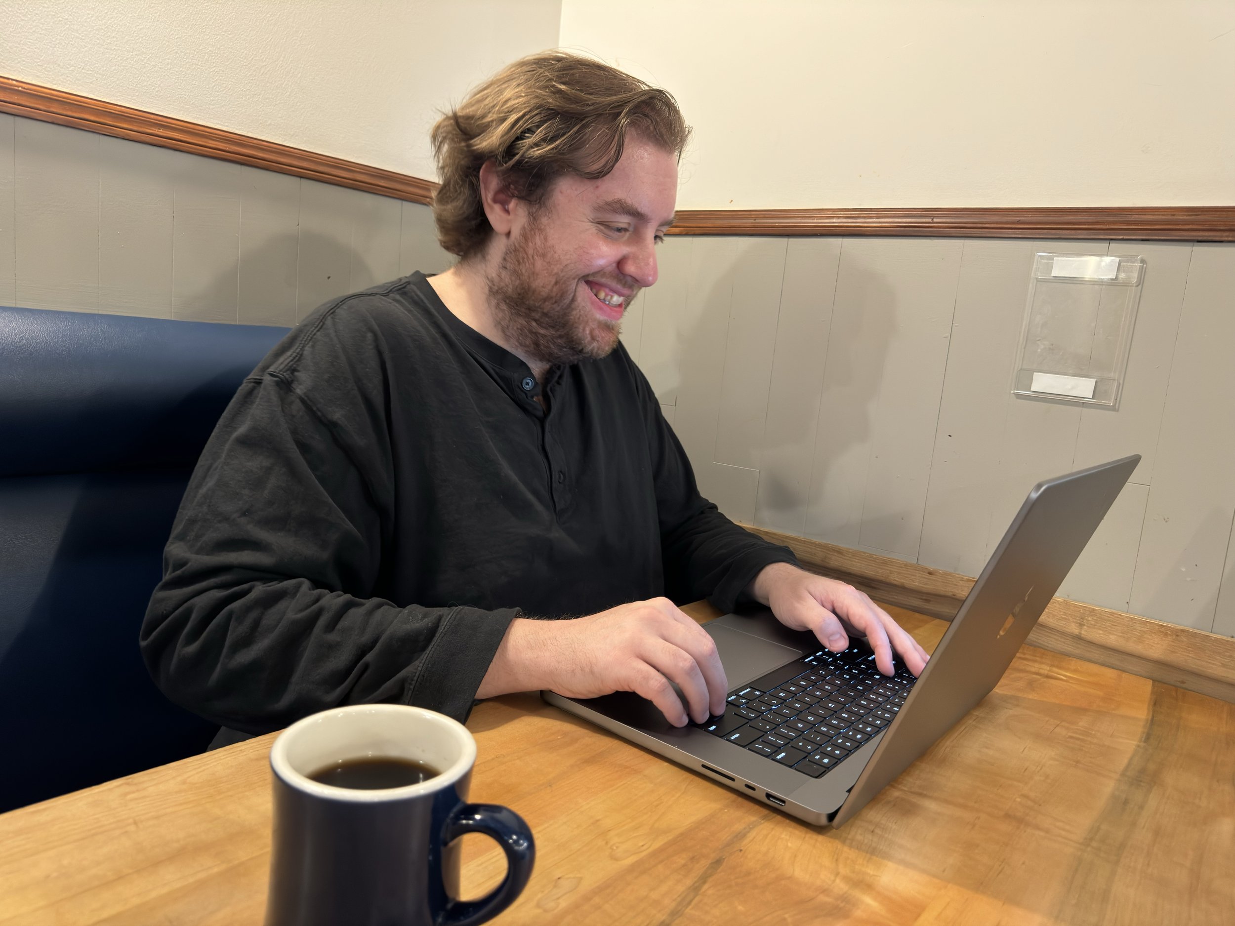  Alex sitting at a table smiling at a MacBook Pro.  A mug of coffee sits in the foreground on the table. 