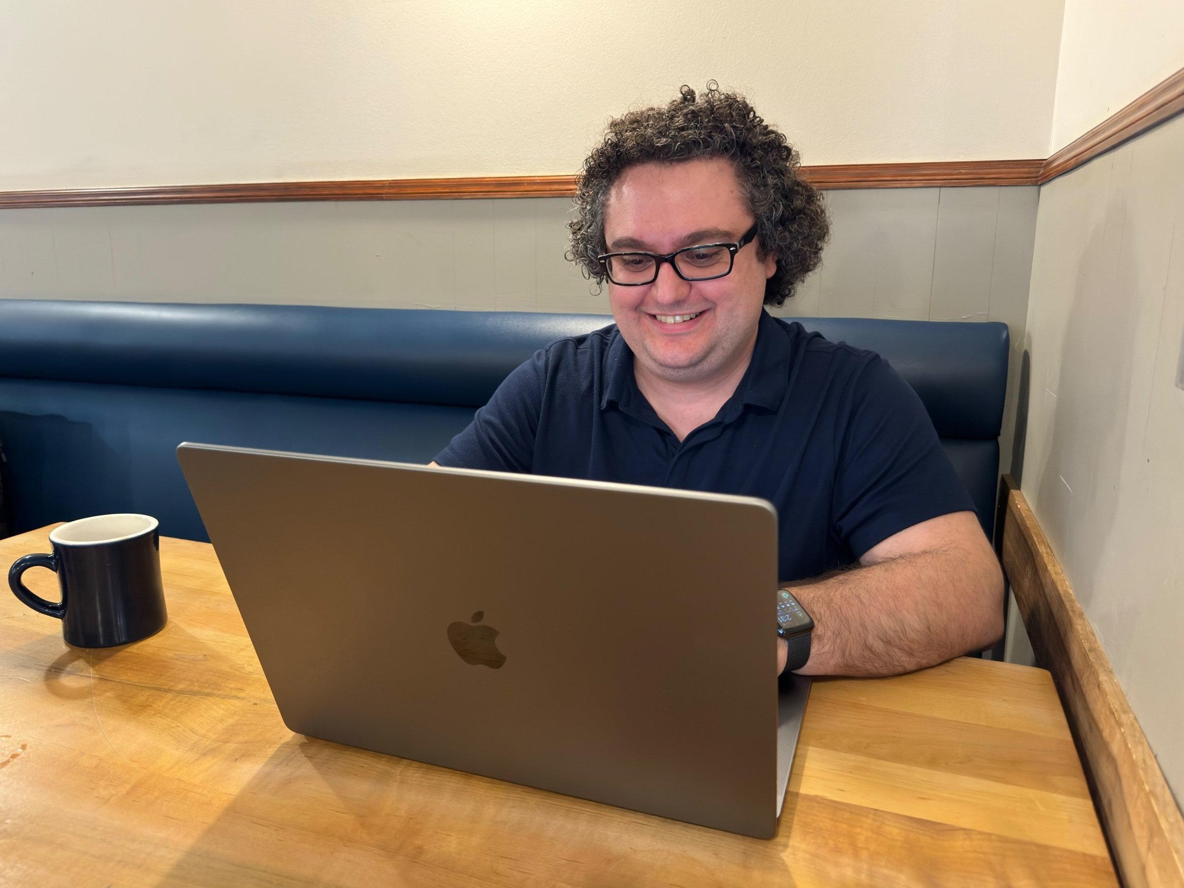  Tony sitting at a table smiling at a MacBook Pro.  A mug of coffee sits next to him on the table. 