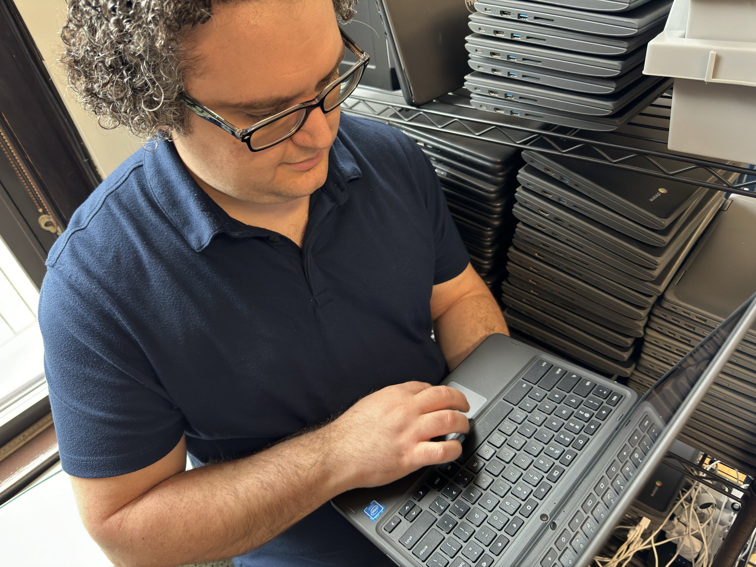  Tony stands holding a Chromebook and looks at the screen.  There are stacks of many other Chromebooks next to him. 