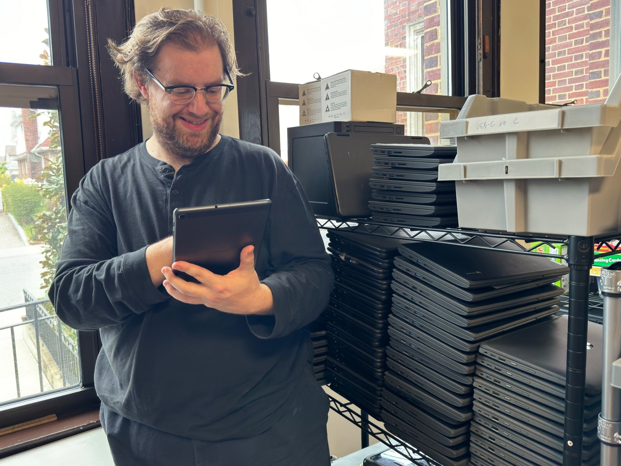  Alex holds an Android tablet, smiling.  He stands next to stacks of many Chromebooks. 