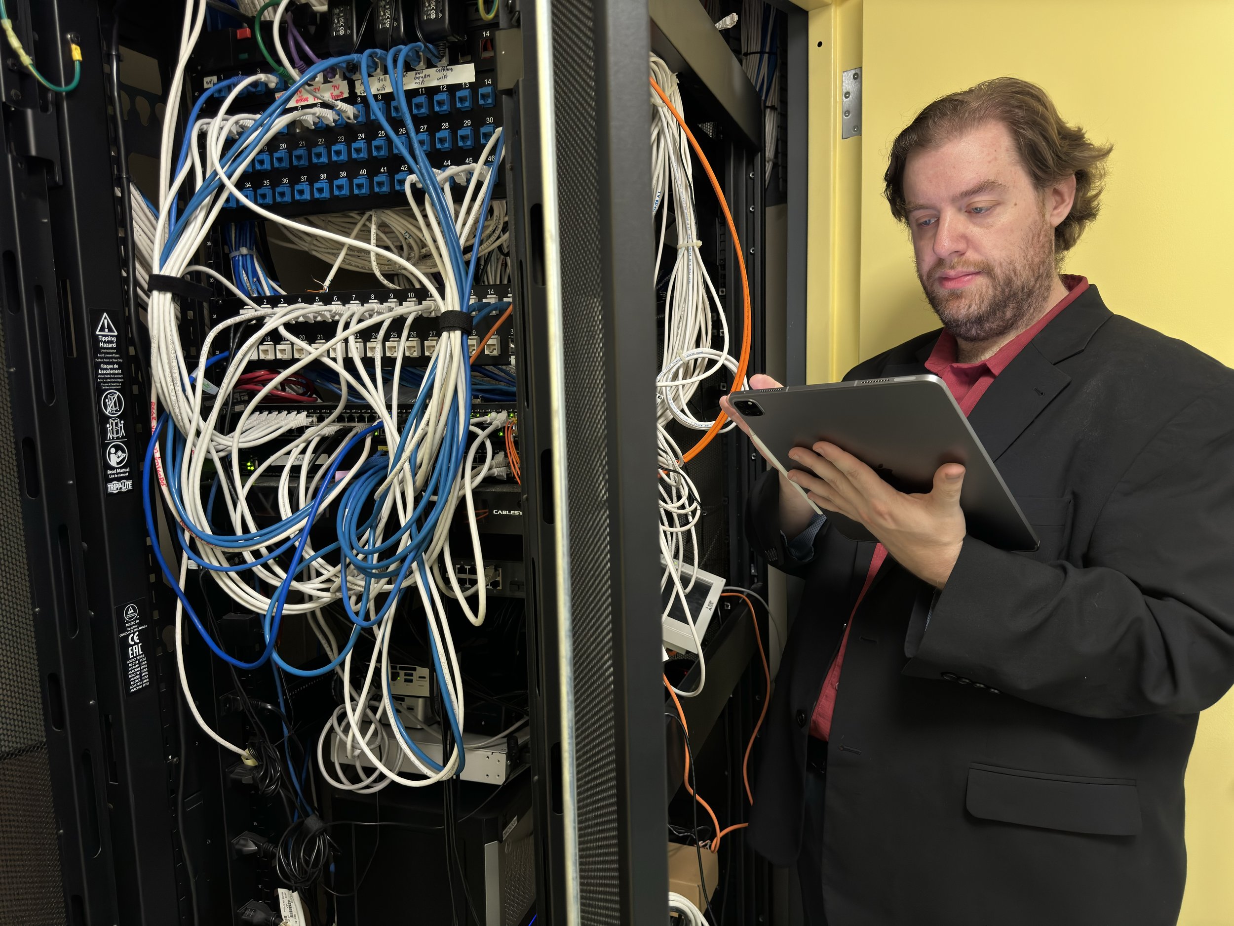  Alex stands in a network closet working on an iPad Pro. 