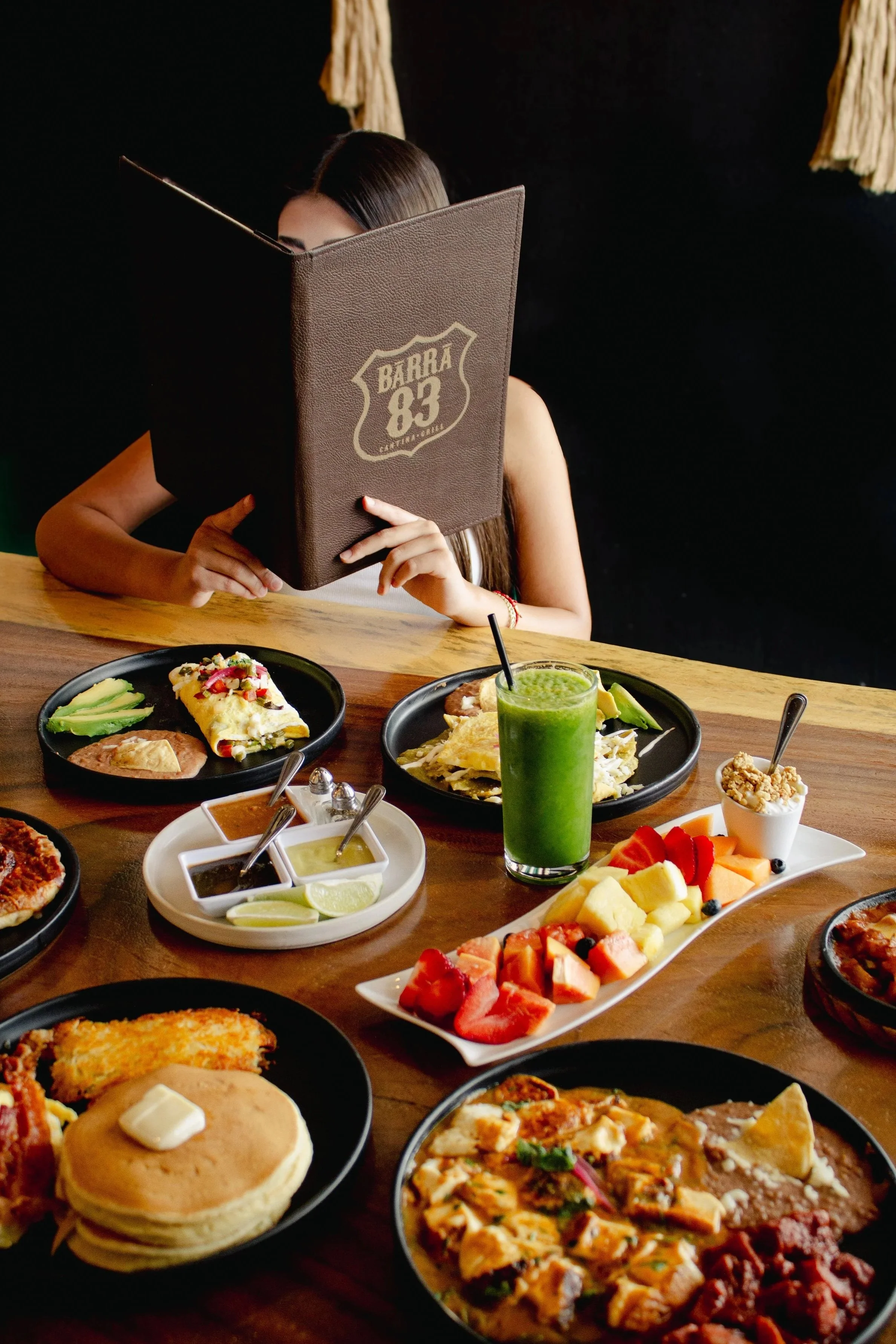 A woman is sitting at a table with Mexican food dishes, including tacos, quesadillas, guacamole, and a fruit platter, while reading a menu in a restaurant.