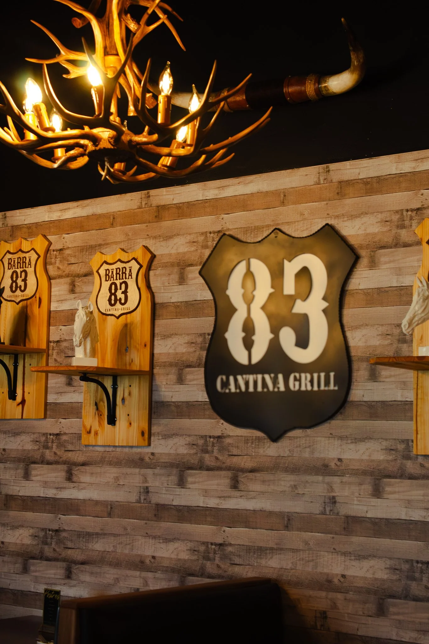 Decorative sign with the number 83 and the words Cantina Grill on a wooden wall, with a chandelier made of antlers and lights hanging above.