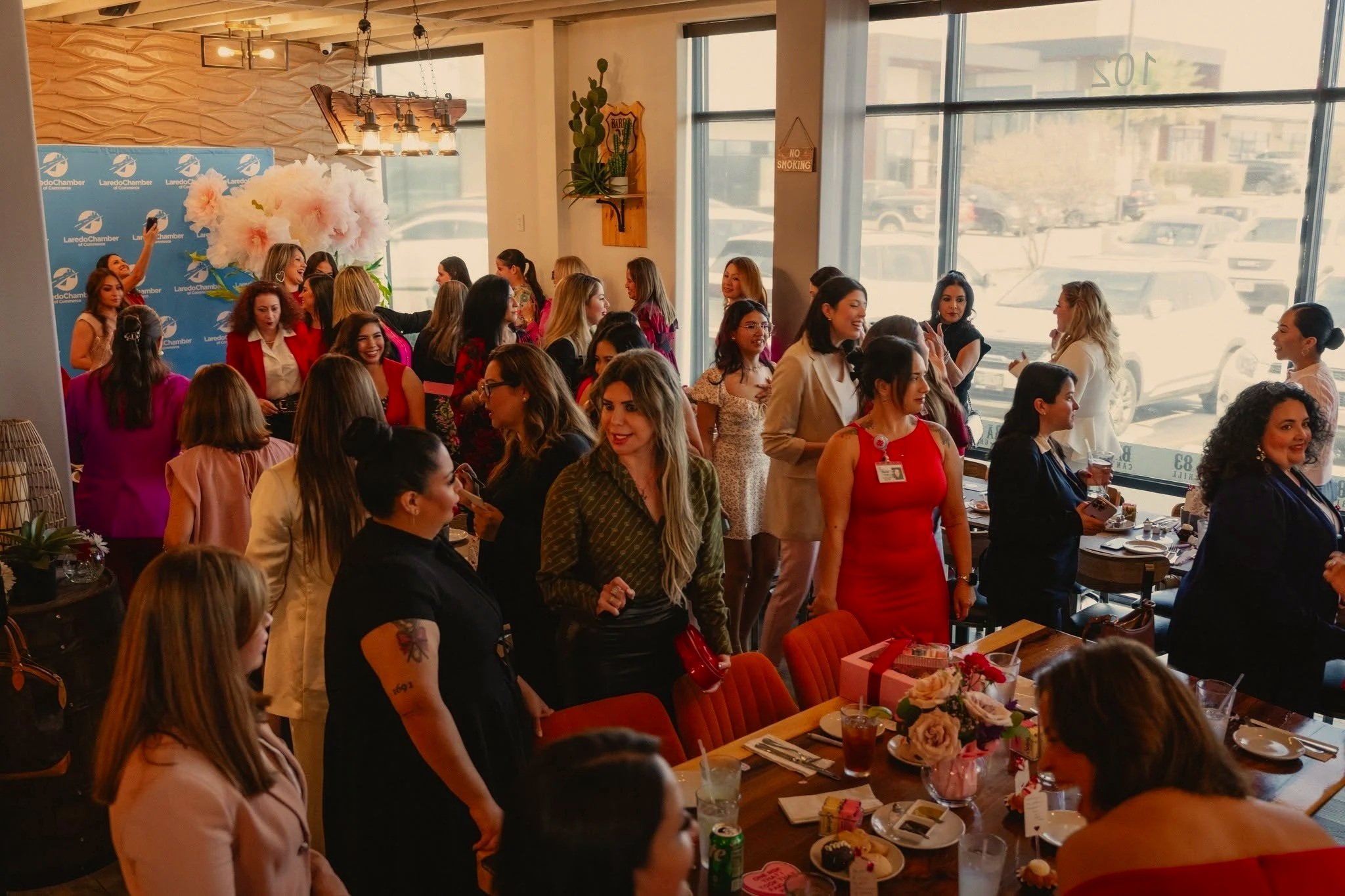 A large group of women gathered inside a restaurant or event space, celebrating together, with some standing and talking, others seated at tables with desserts and drinks, and a floral backdrop with the logo of Laredo Chamber in the background.