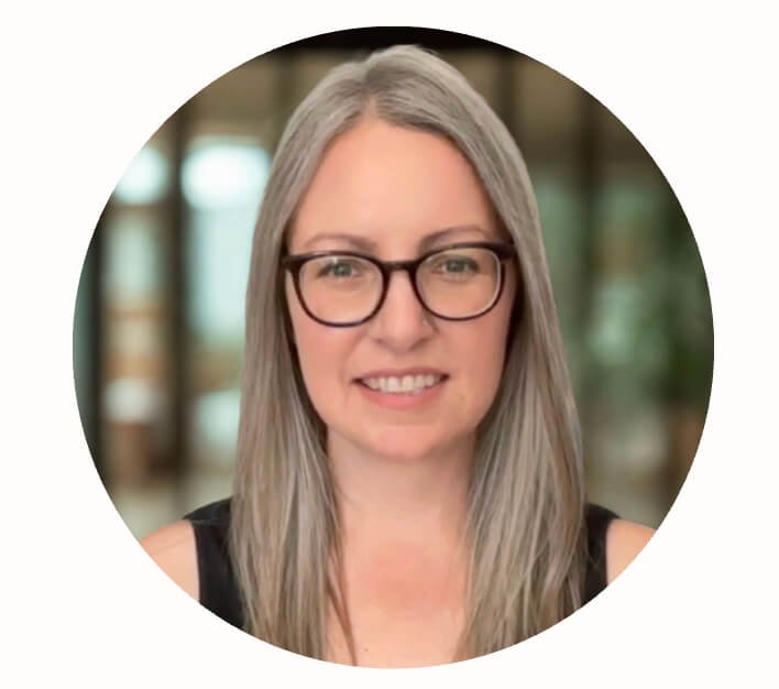 Portrait of a woman with long gray hair, wearing glasses and a black sleeveless top, smiling indoors with blurred background.